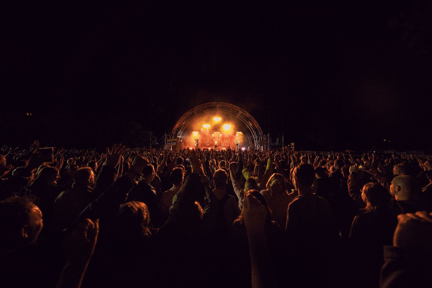 Large outdoor concert crowd at night with hands raised toward a glowing stage with performers under an arch.
