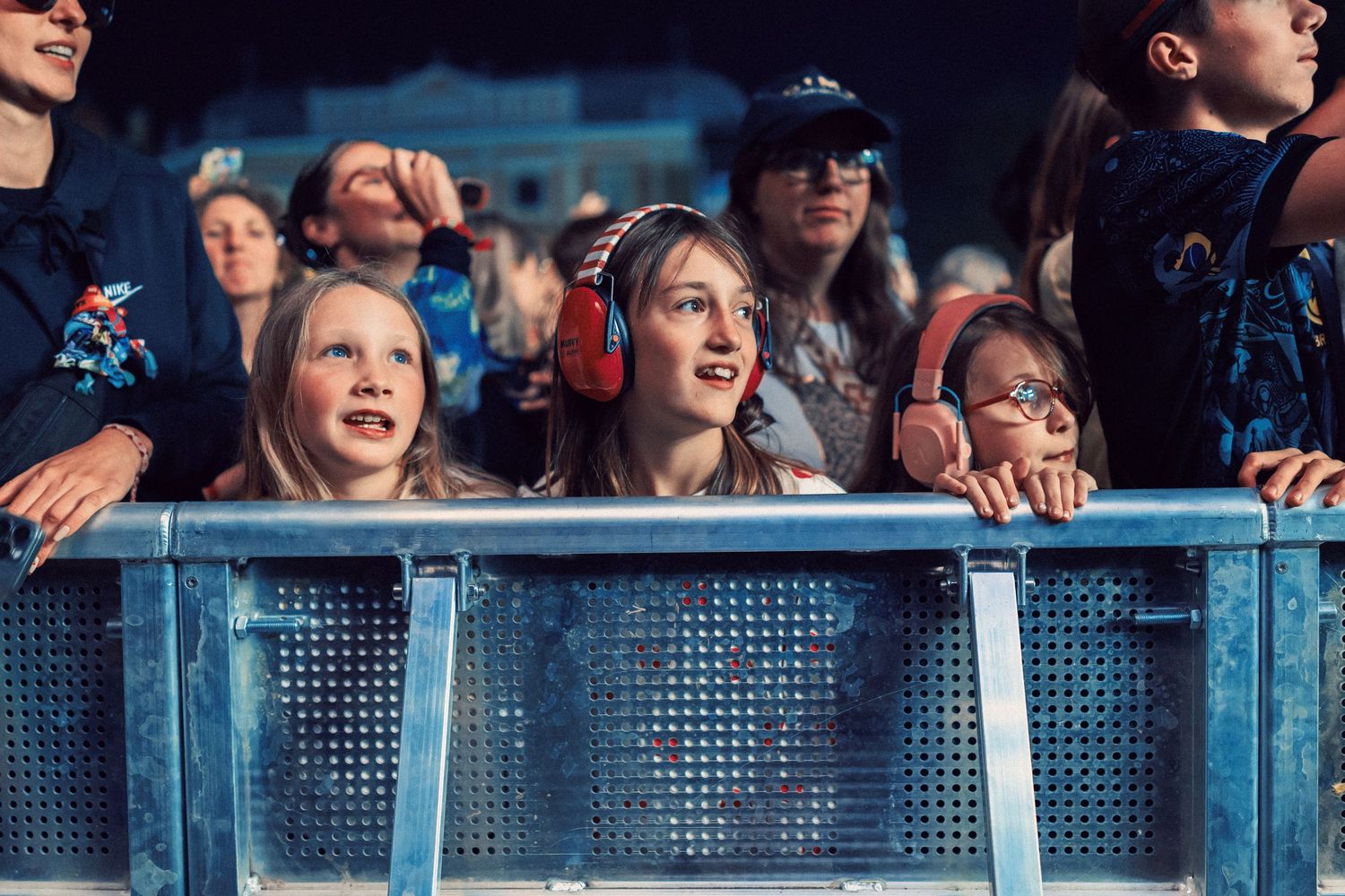 Children wearing ear protection smile at a live music festival, leaning on a metal barrier in a crowd.