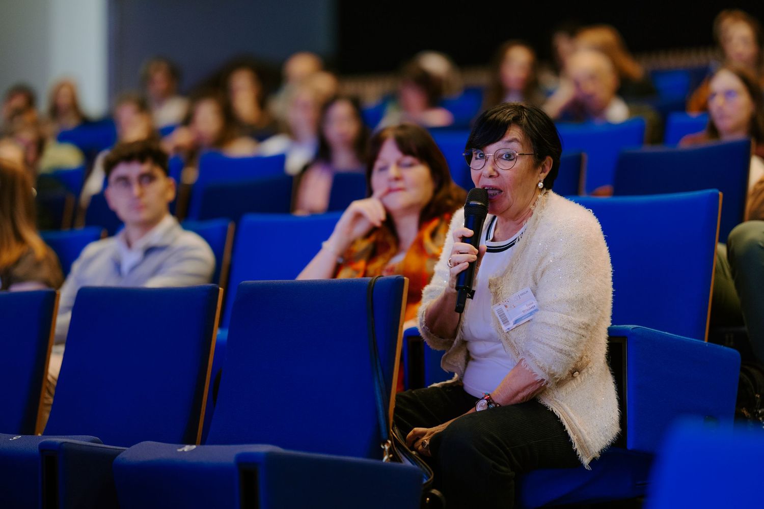 Woman speaking into microphone at a conference seated among audience members in blue chairs.