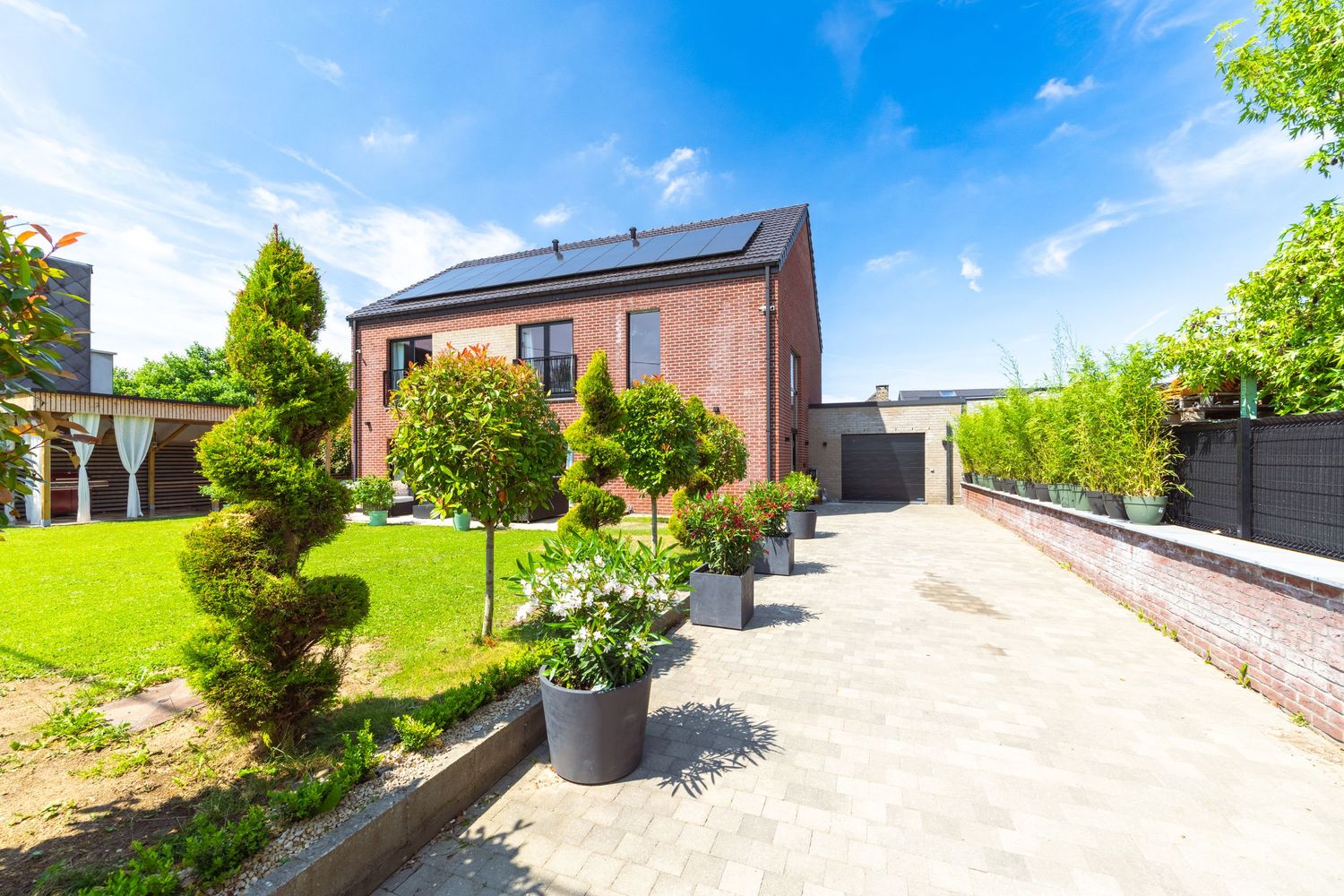 Brick house with solar panels, manicured garden, potted trees, and paved driveway under a bright blue sky.