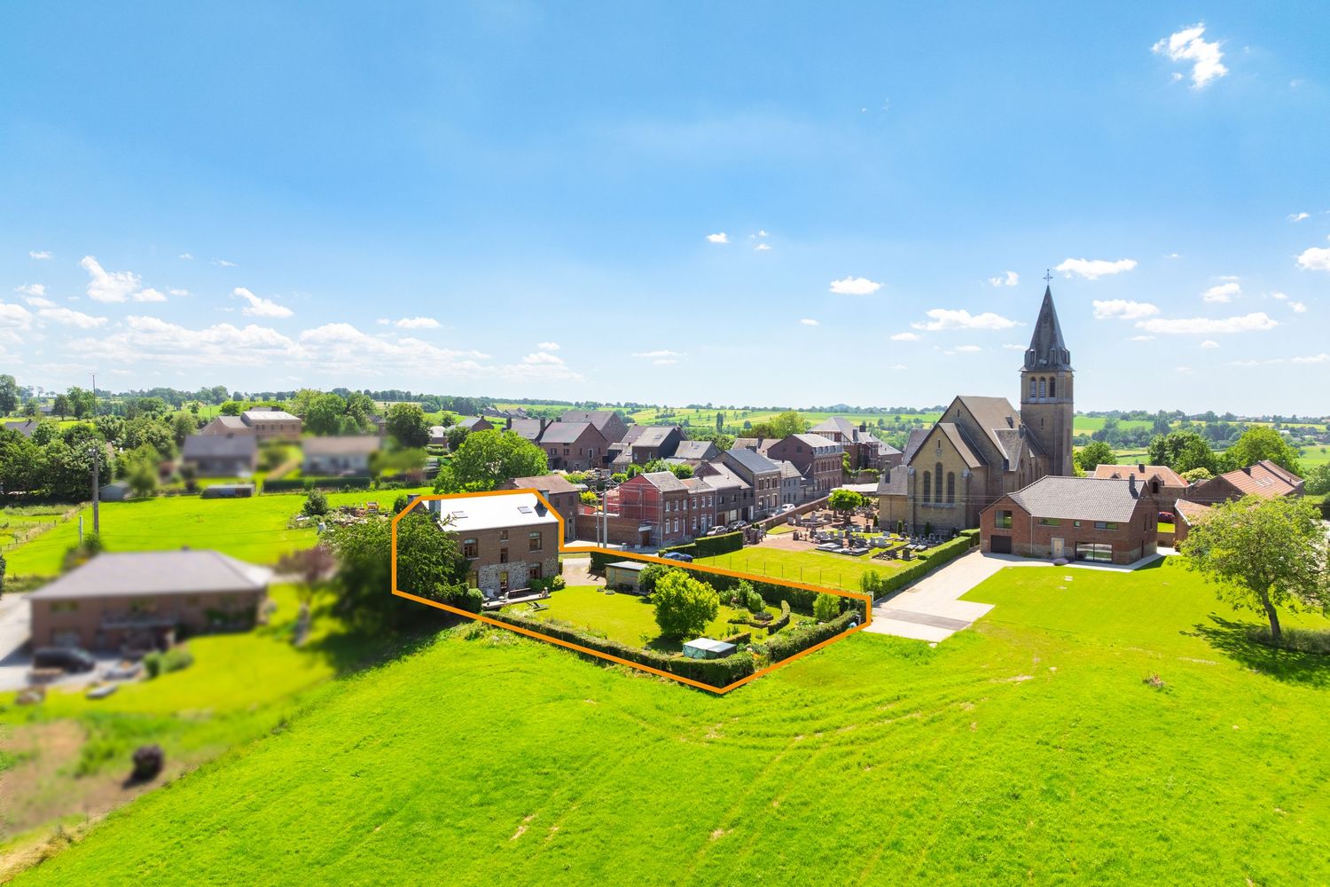 Aerial drone view of a charming Belgian village with a historic church steeple surrounded by lush green fields.
