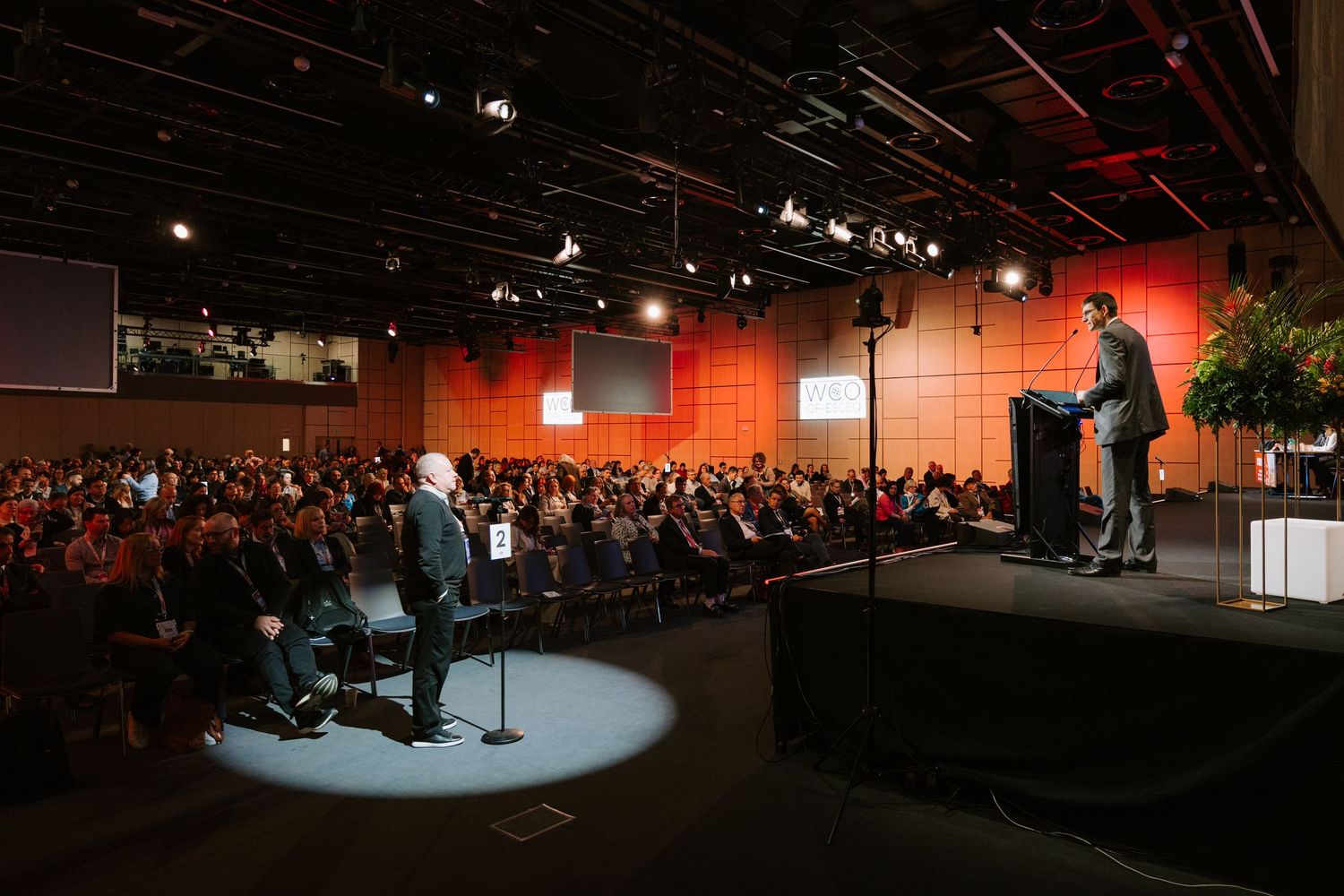A speaker presents on stage to a large audience in a modern conference hall with orange accent walls.
