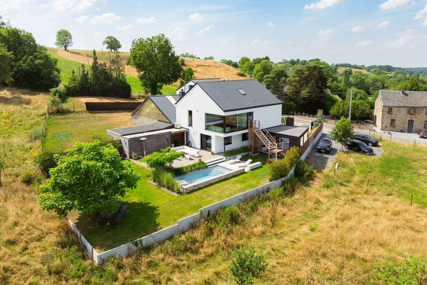 Aerial view of a modern white house with a swimming pool, lush garden, and scenic countryside surroundings.