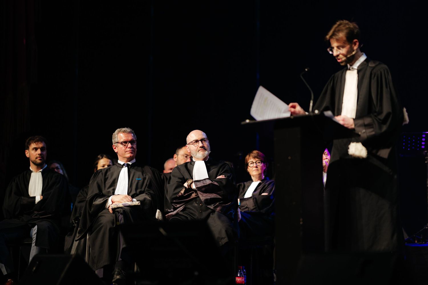 Academic ceremony with robed speakers at podium on darkened theater stage.