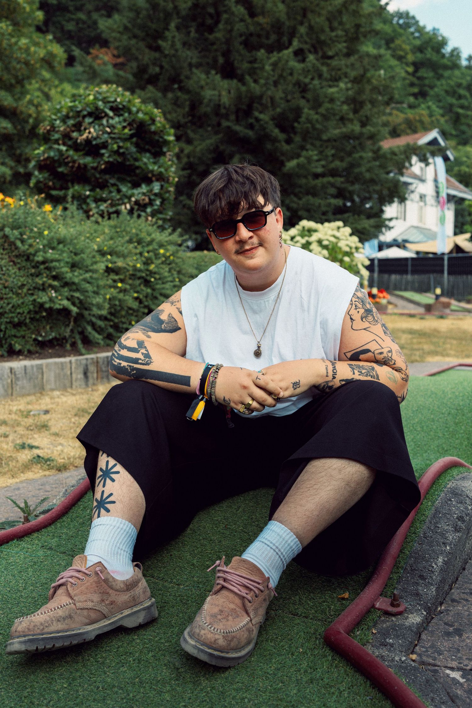 Tattooed person in sunglasses, white sleeveless shirt, black shorts, sitting on mini golf course turf outdoors.