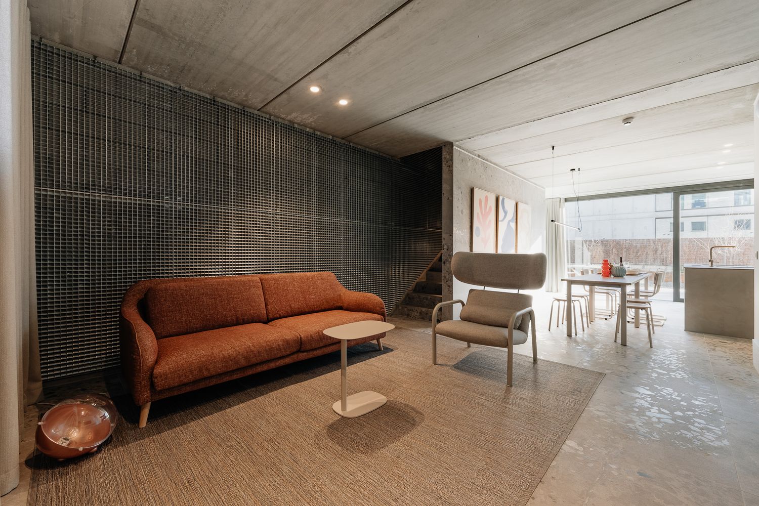 Contemporary living room with rust-colored sofa, dark grid feature wall, concrete ceiling, and open dining area.