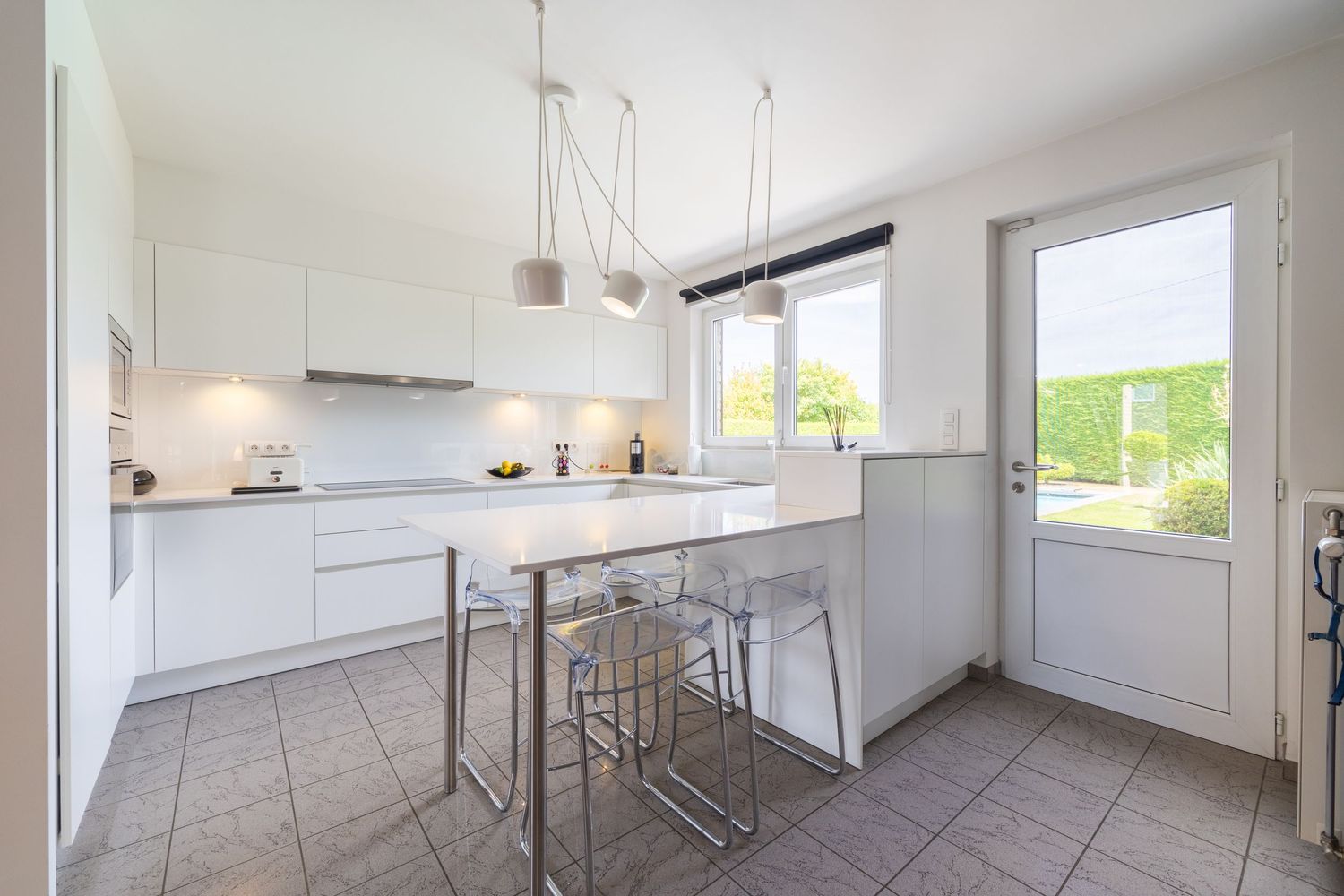 Modern white kitchen with island, bar stools, pendant lights, and a glass door leading to a garden.