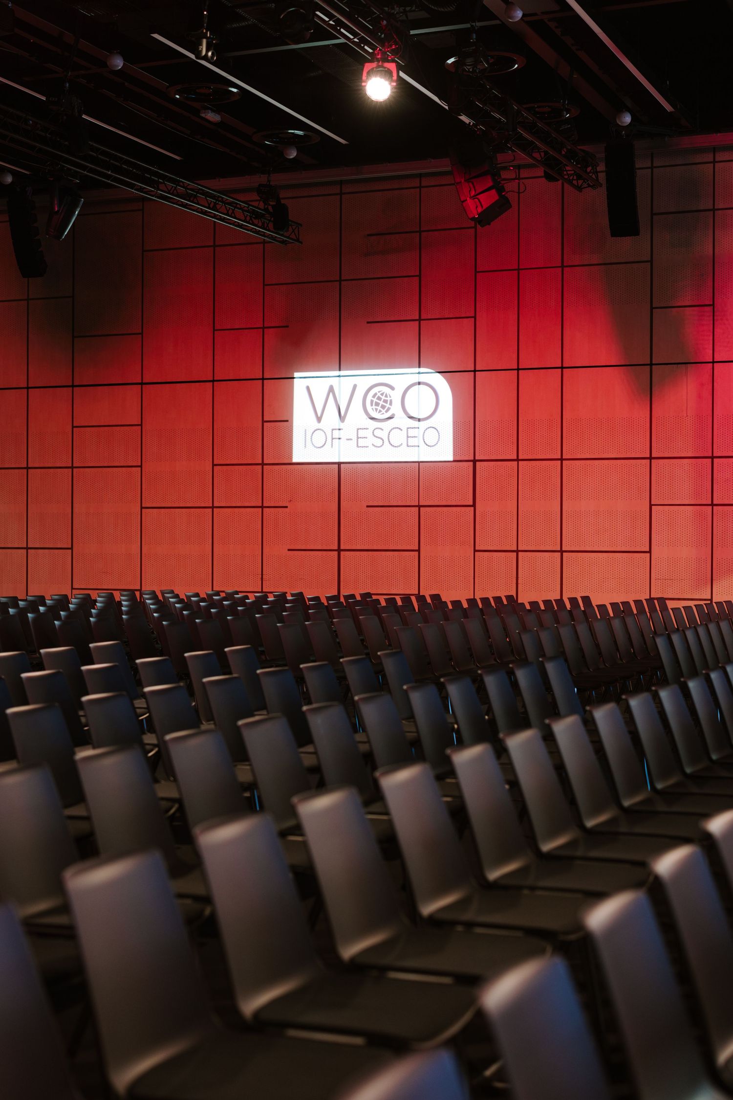 Empty auditorium with rows of black chairs facing a red wall displaying the WCO ESCEO logo on stage.