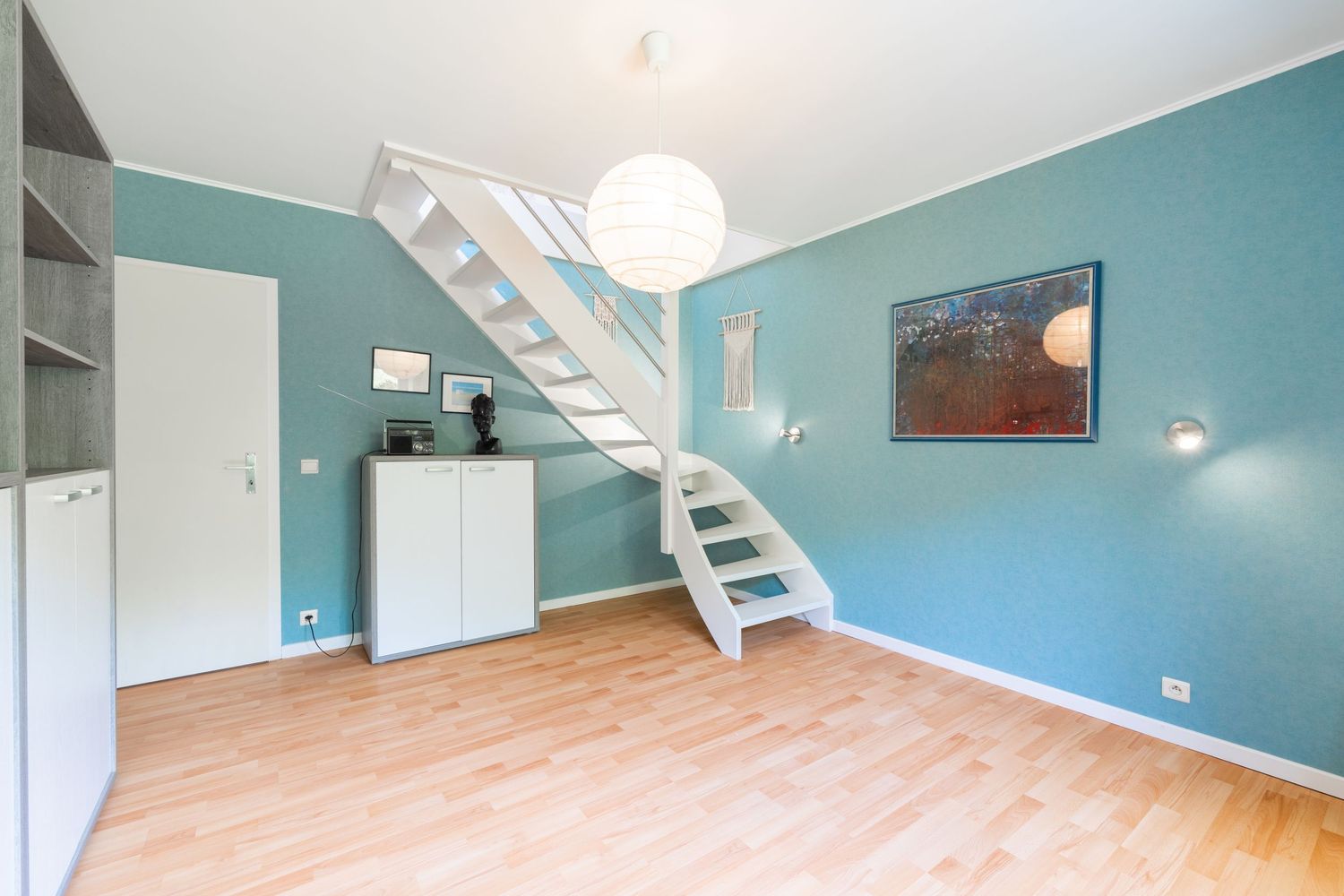 Bright living room with teal walls, white staircase, hardwood floors, and minimalist decor including a white cabinet and pendant light.