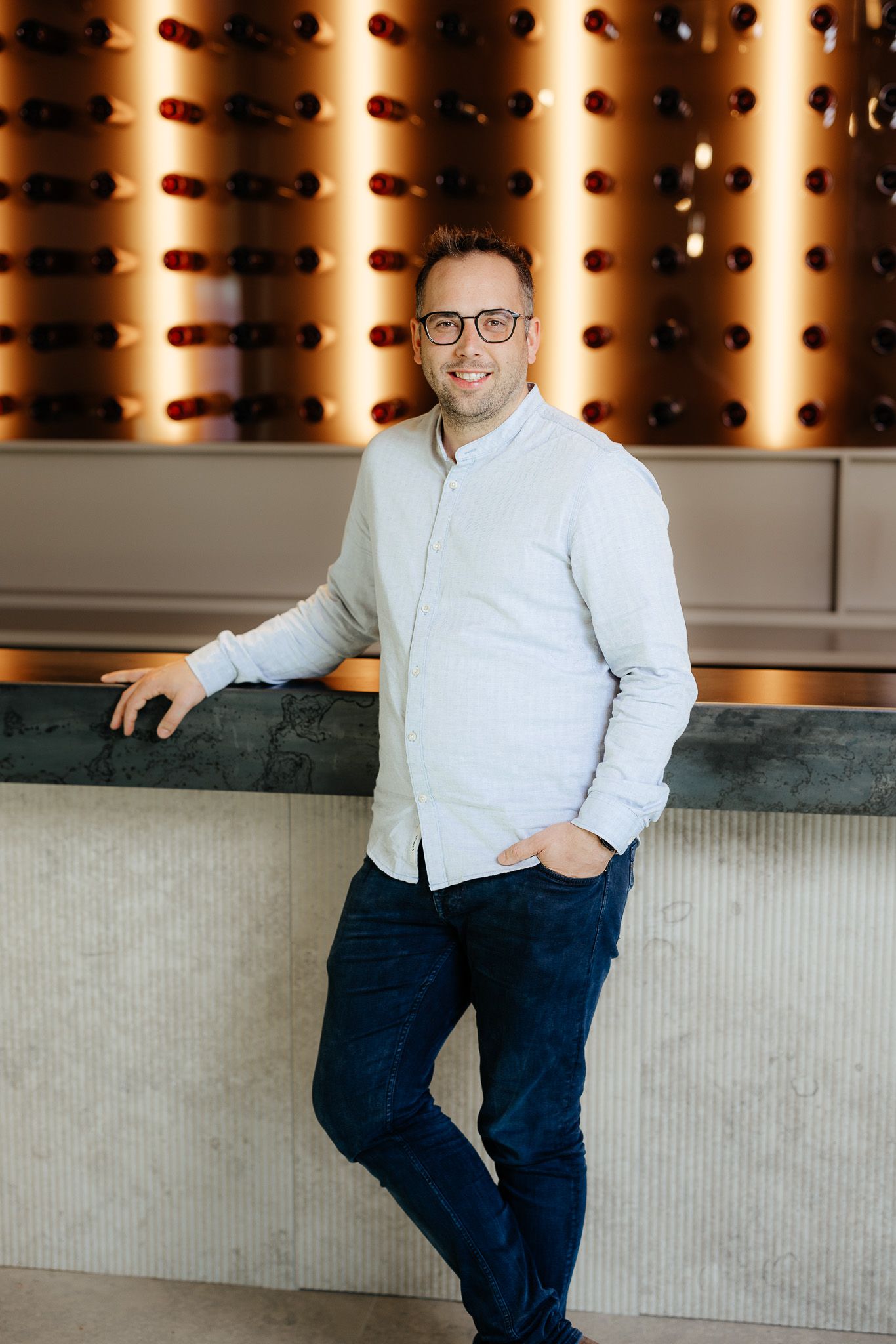 Smiling man in glasses and light shirt leaning on counter in front of illuminated wine rack.