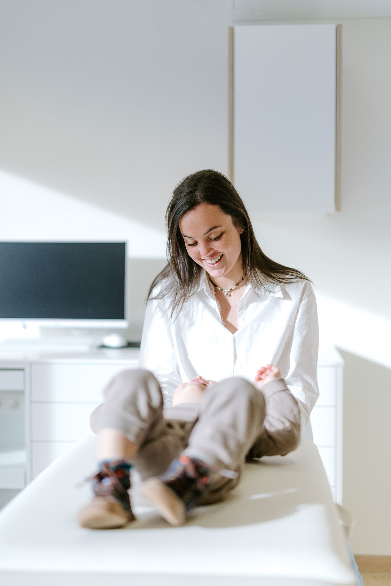 Smiling woman in white shirt sits cross-legged on desk in bright modern home office space.