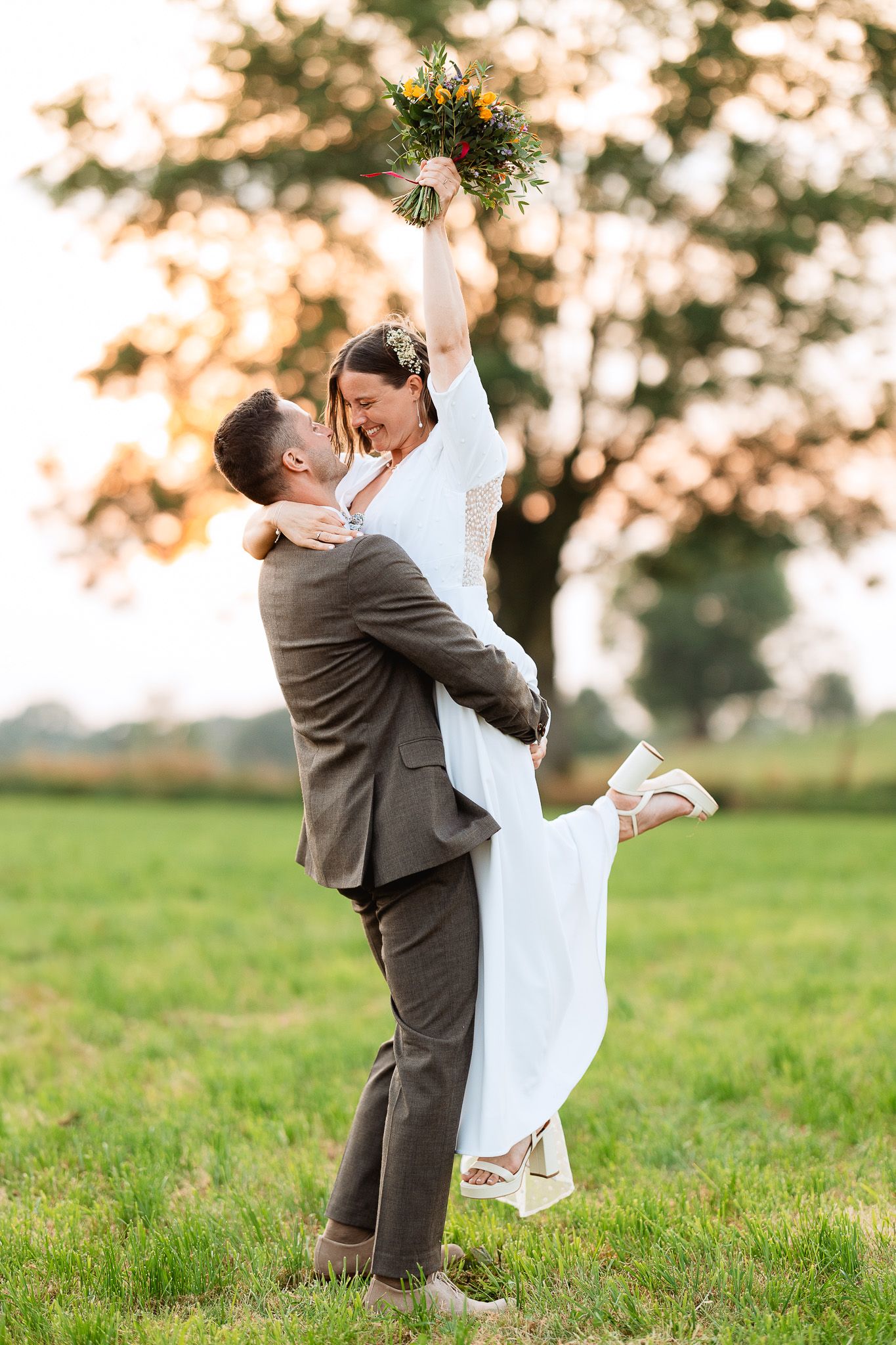 Groom lifts bride in white dress as she raises colorful bouquet triumphantly in a sunny green field.