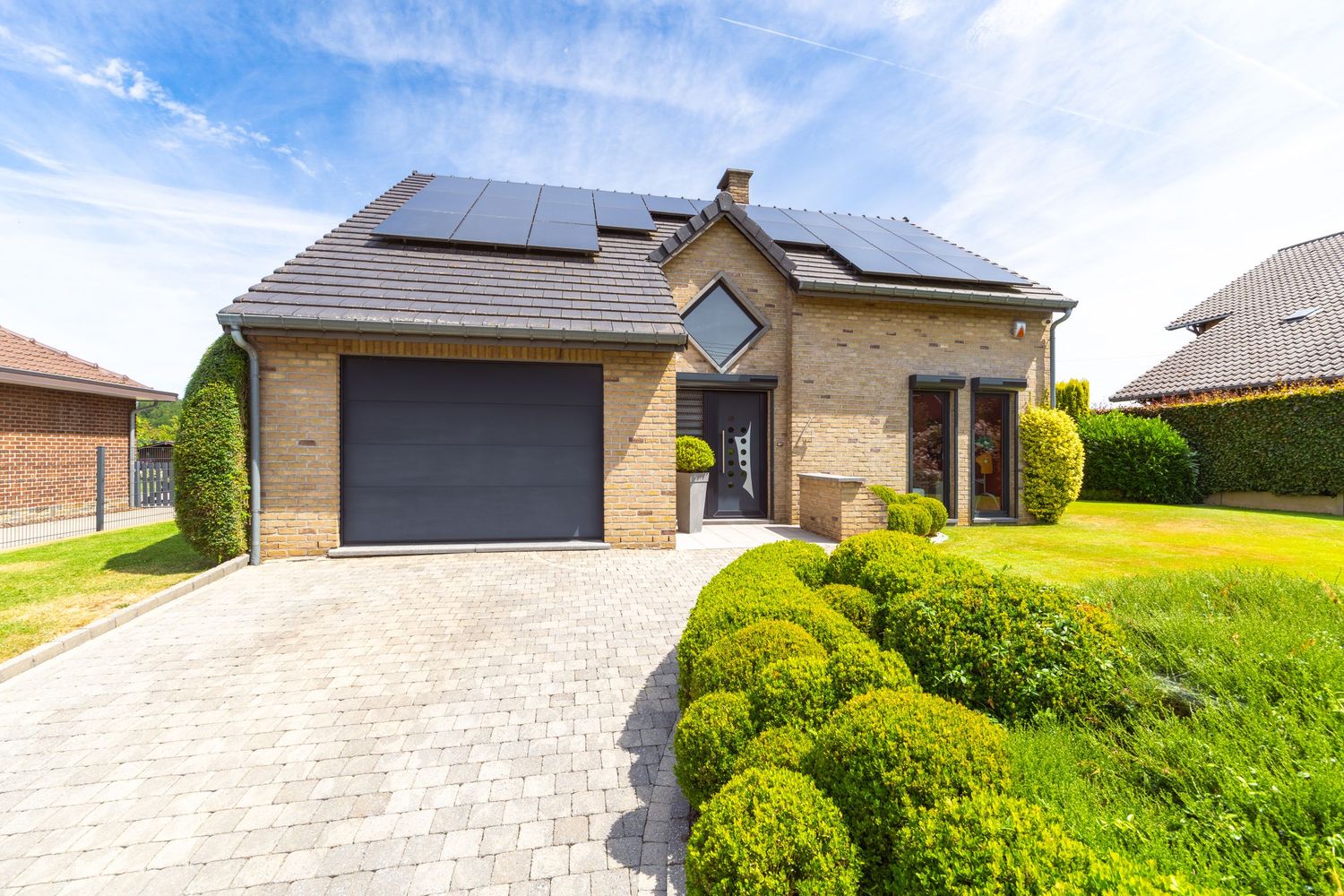 Brick house with solar panels, black garage door, manicured hedges, and paved driveway on a sunny day.