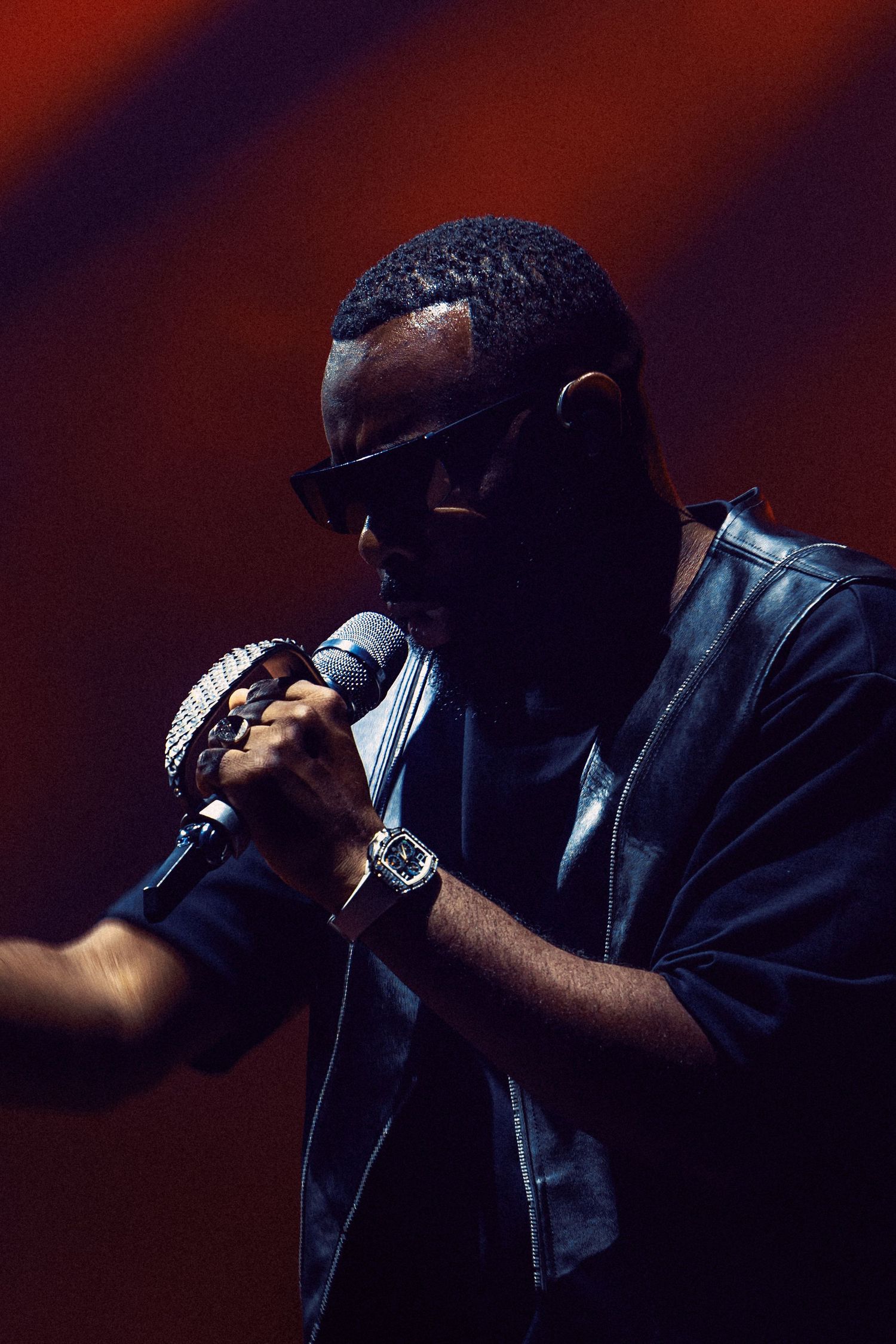 A male performer wearing sunglasses and a leather jacket sings into a microphone on a dramatic red-lit stage.