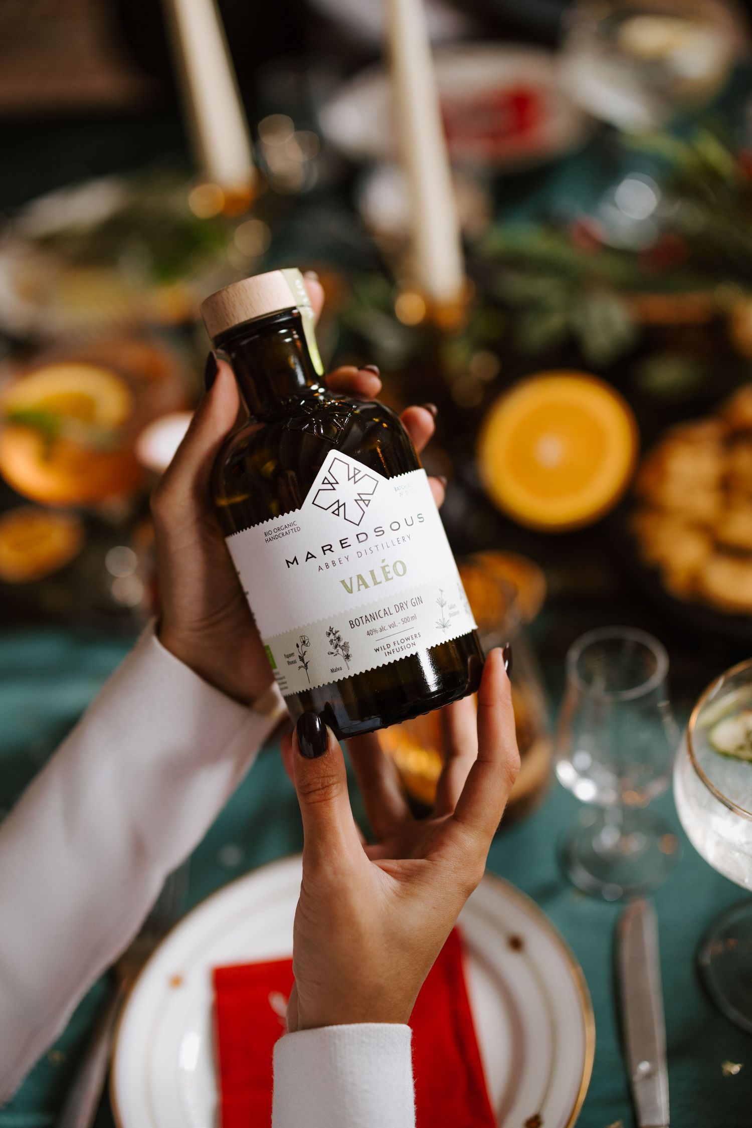 Two hands holding a Maredsous Valeo bottle over a decorated holiday dining table with oranges and candles.