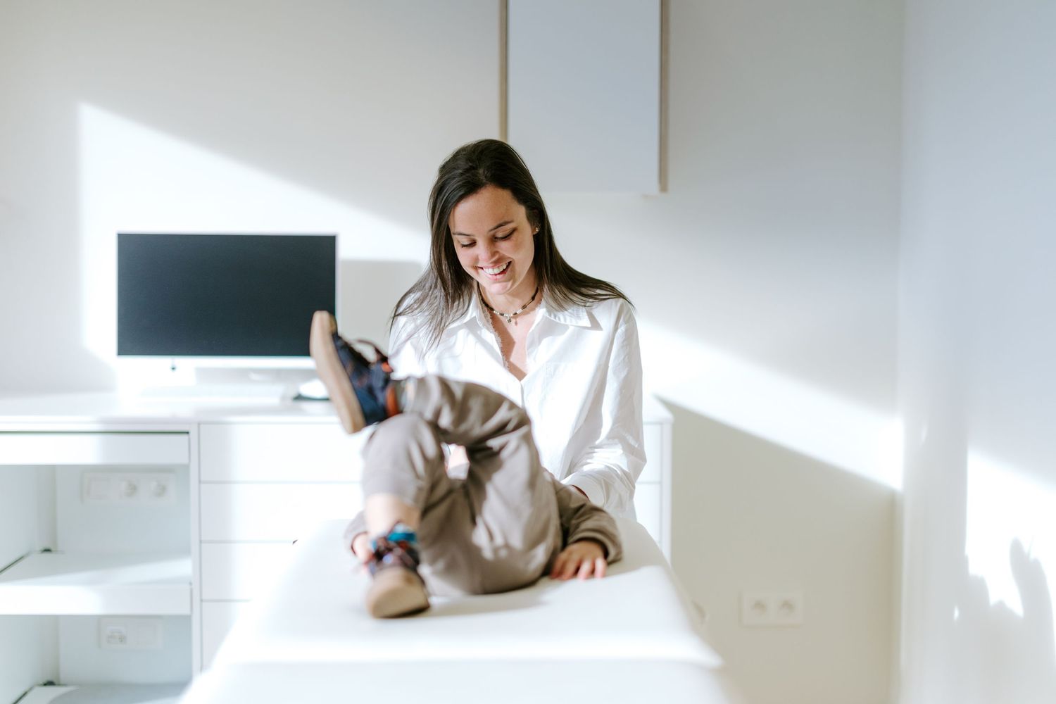 A smiling woman in a white shirt sits cross-legged on a white desk in a bright modern office with a computer monitor.
