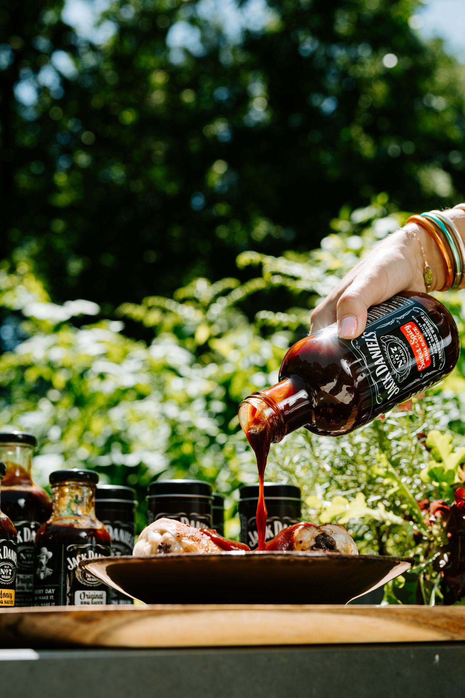 Hand pouring Jack Daniel's BBQ sauce over grilled meat on a wooden board outdoors with sauce bottles in background.