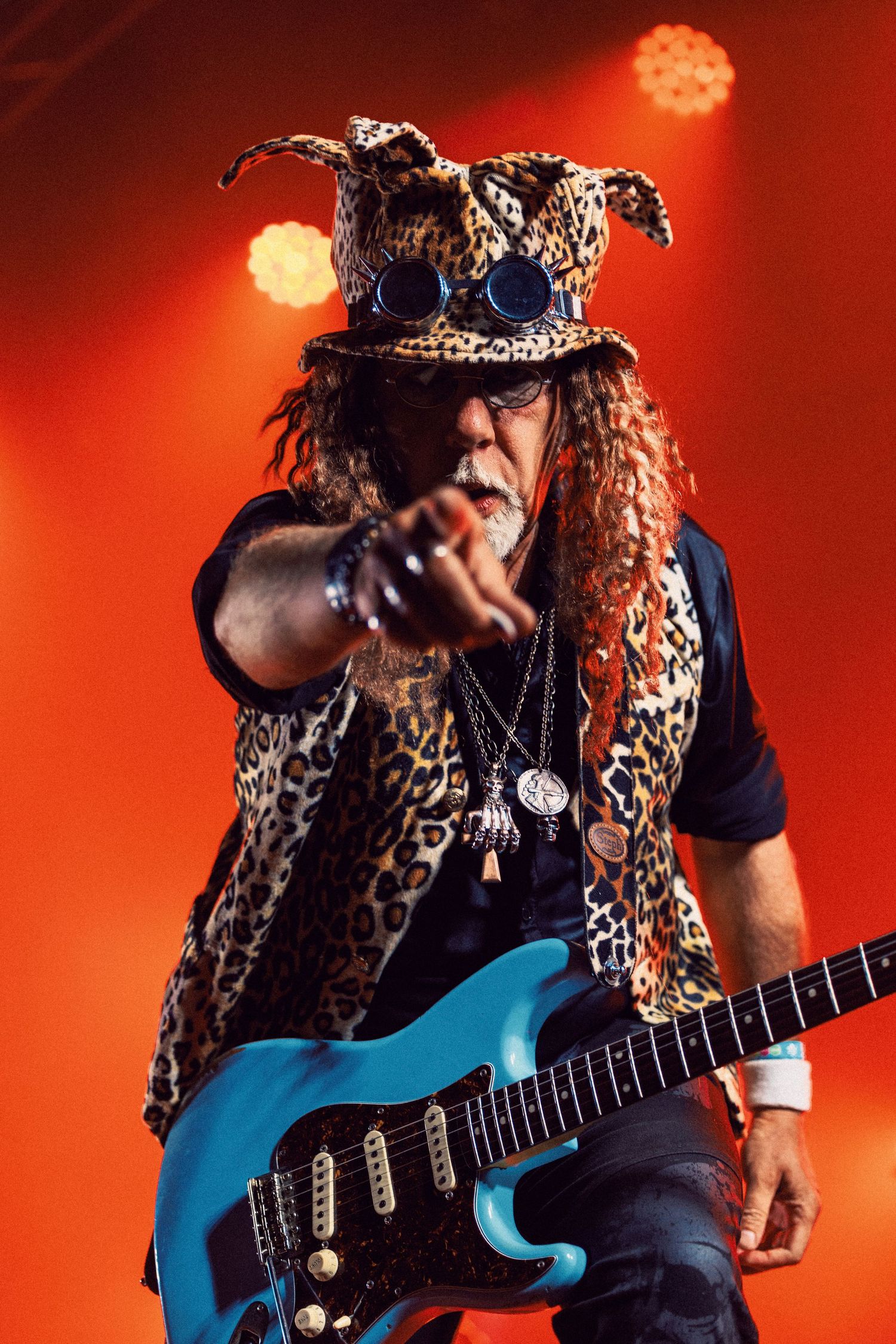 Flamboyant rock guitarist in leopard print outfit and hat playing blue electric guitar on stage with red lighting.