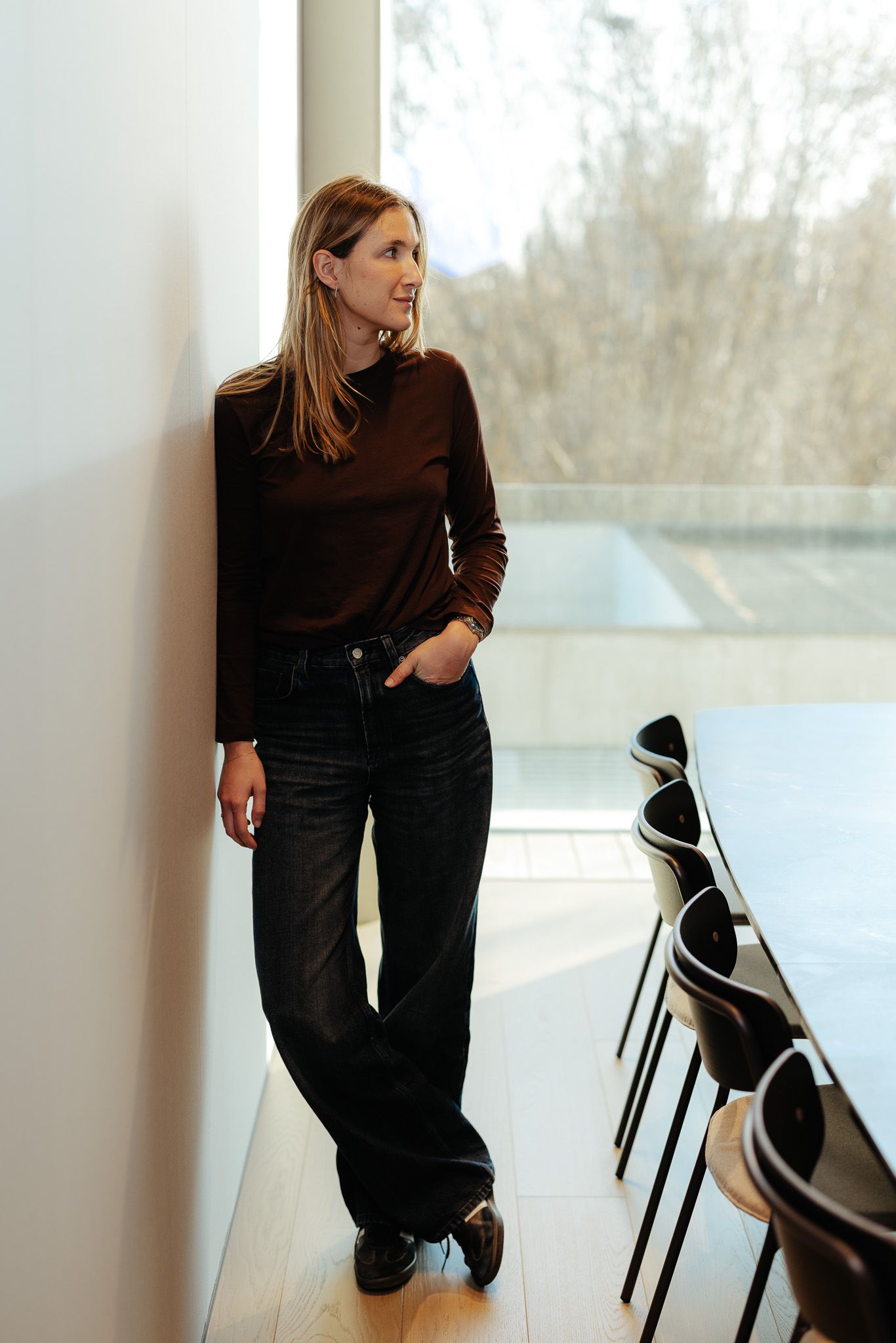 Blonde woman in brown top and black jeans leaning against white wall near large window.