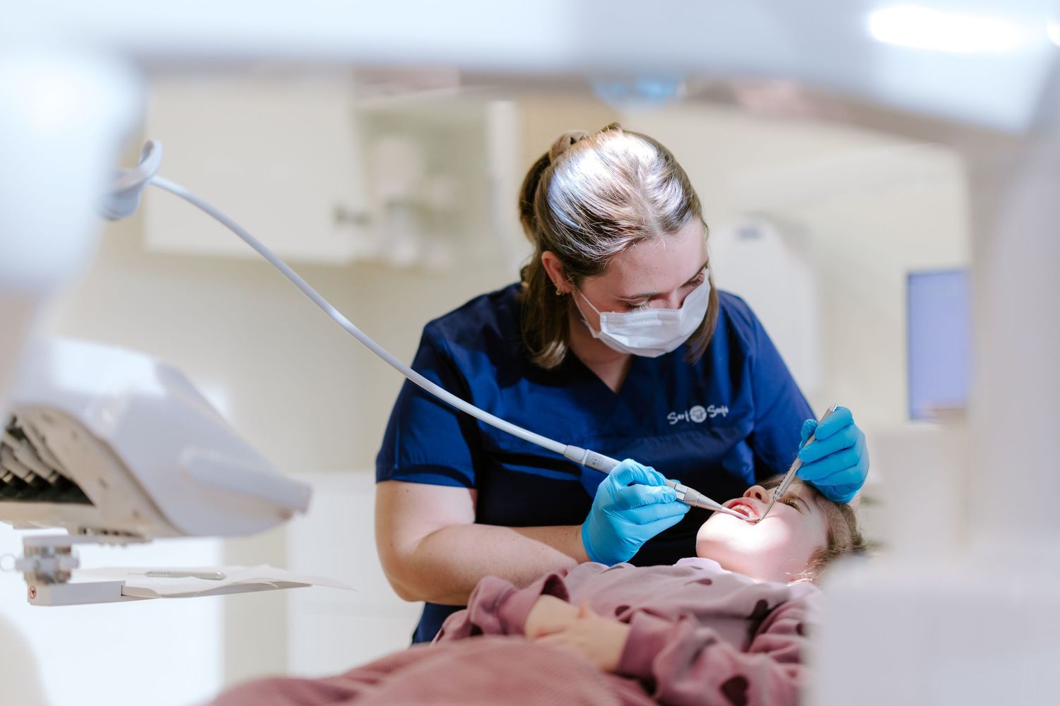 Dental hygienist in blue scrubs and mask performing a dental examination on a patient in a modern clinic.