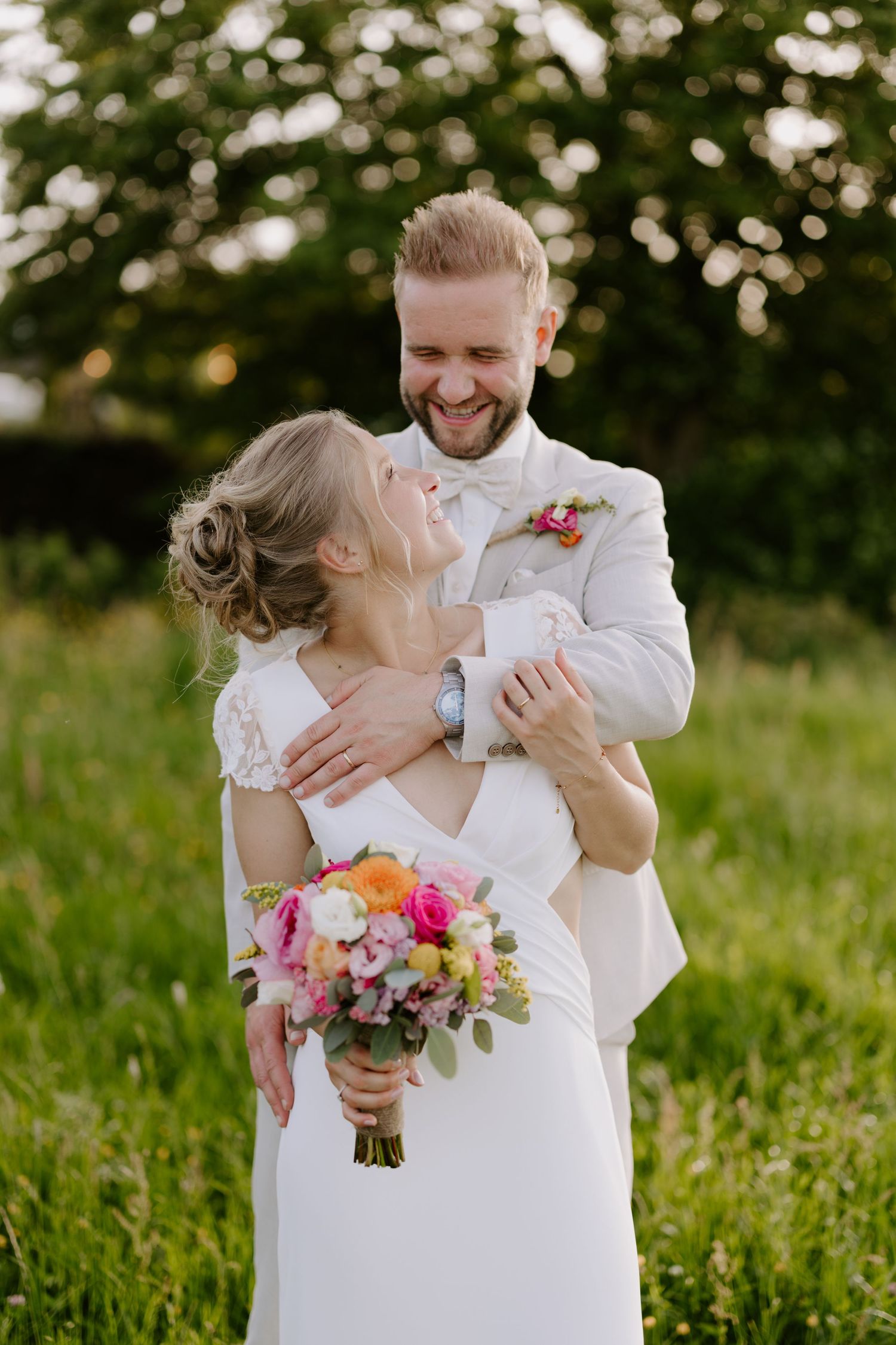 Joyful groom in white suit lifts smiling bride holding colorful bouquet in a sunny green meadow.