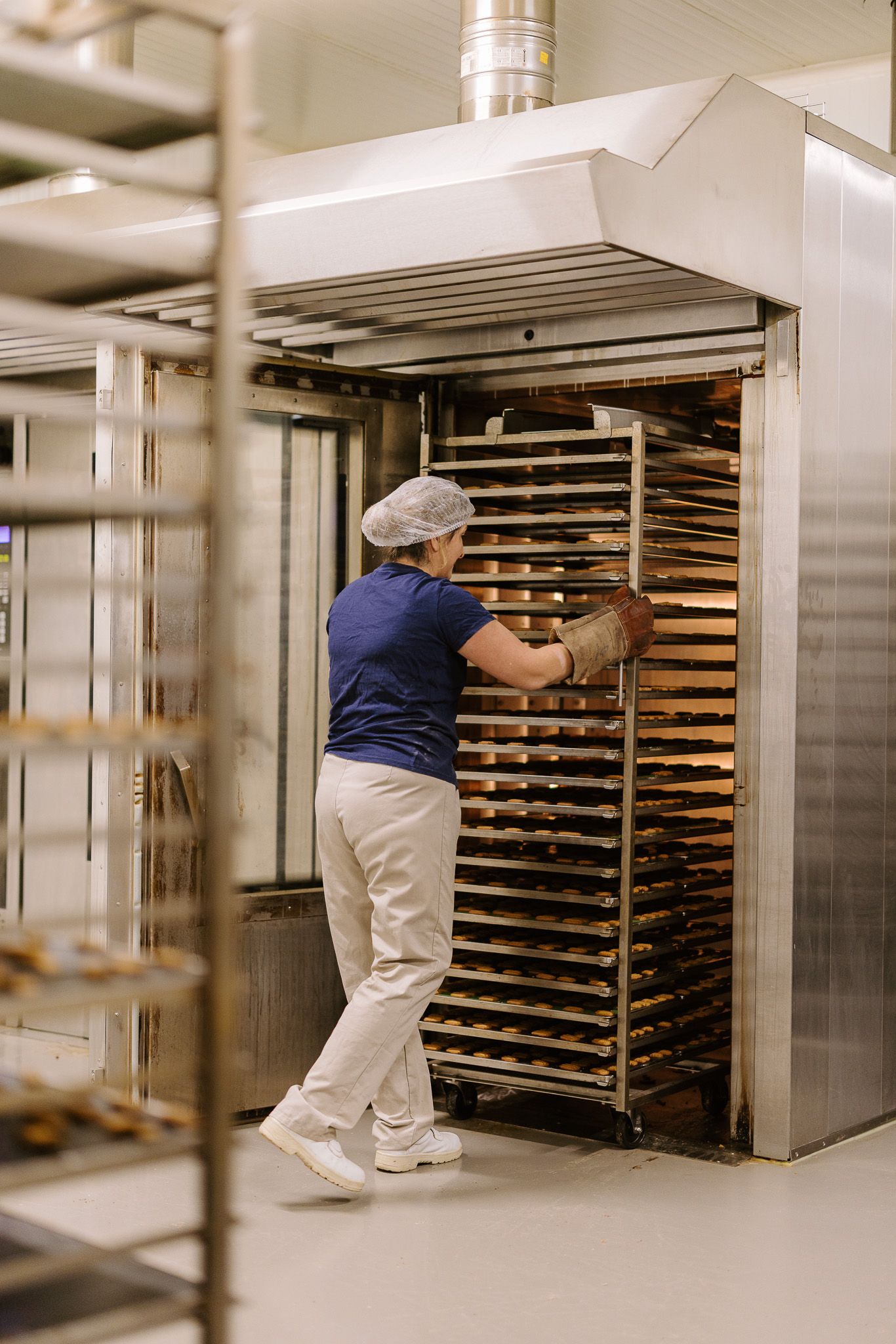Female baker in hairnet loads rack of baked goods into large commercial deck oven in bakery.