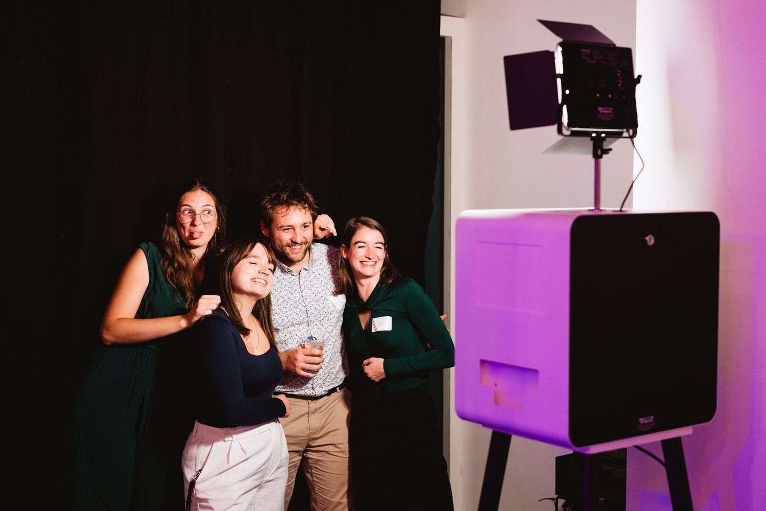Four friends pose and laugh together at a photo booth setup with purple lighting and professional camera equipment.