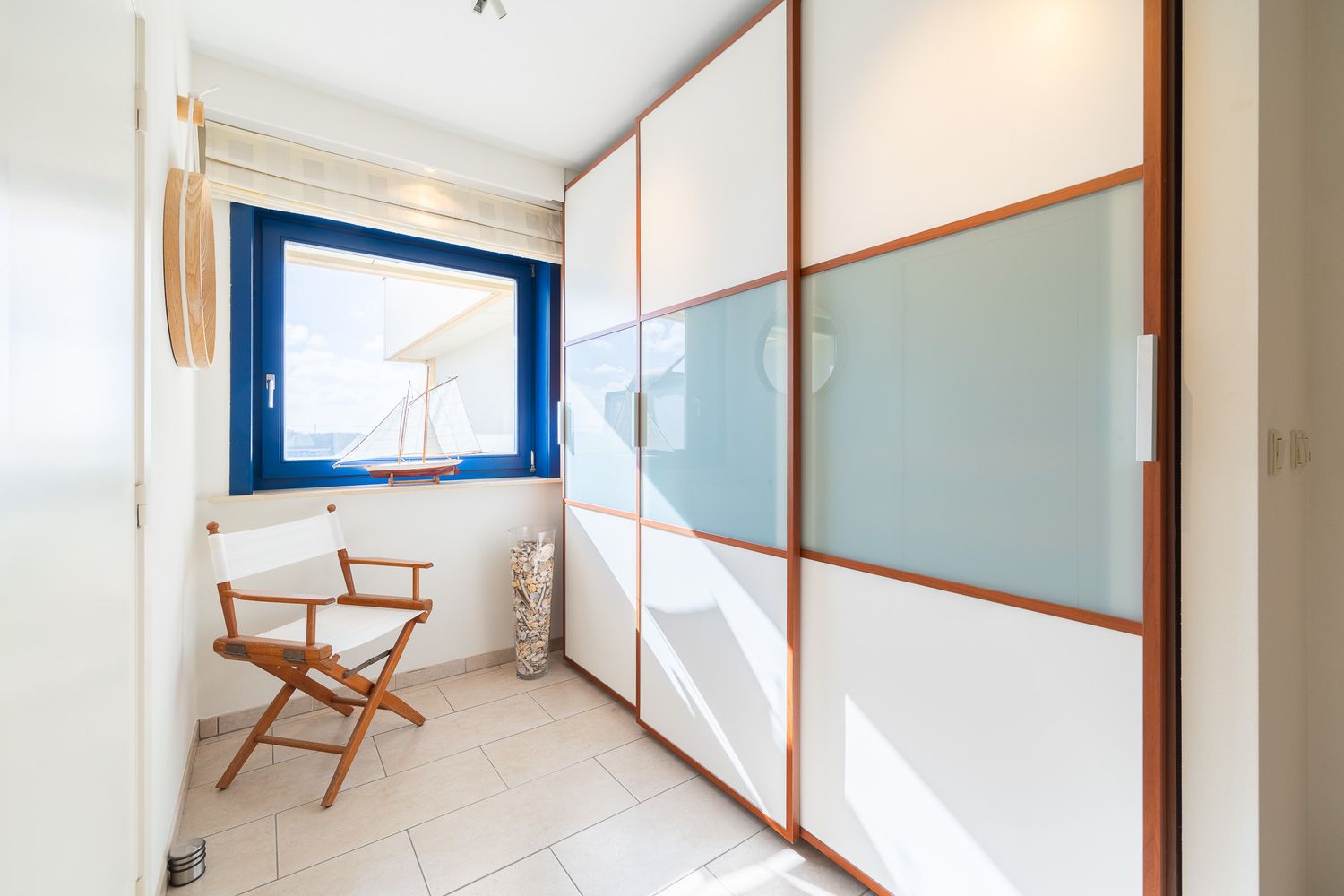 Bright room with sliding wardrobe doors, wooden chair, and blue-framed window with sea view.