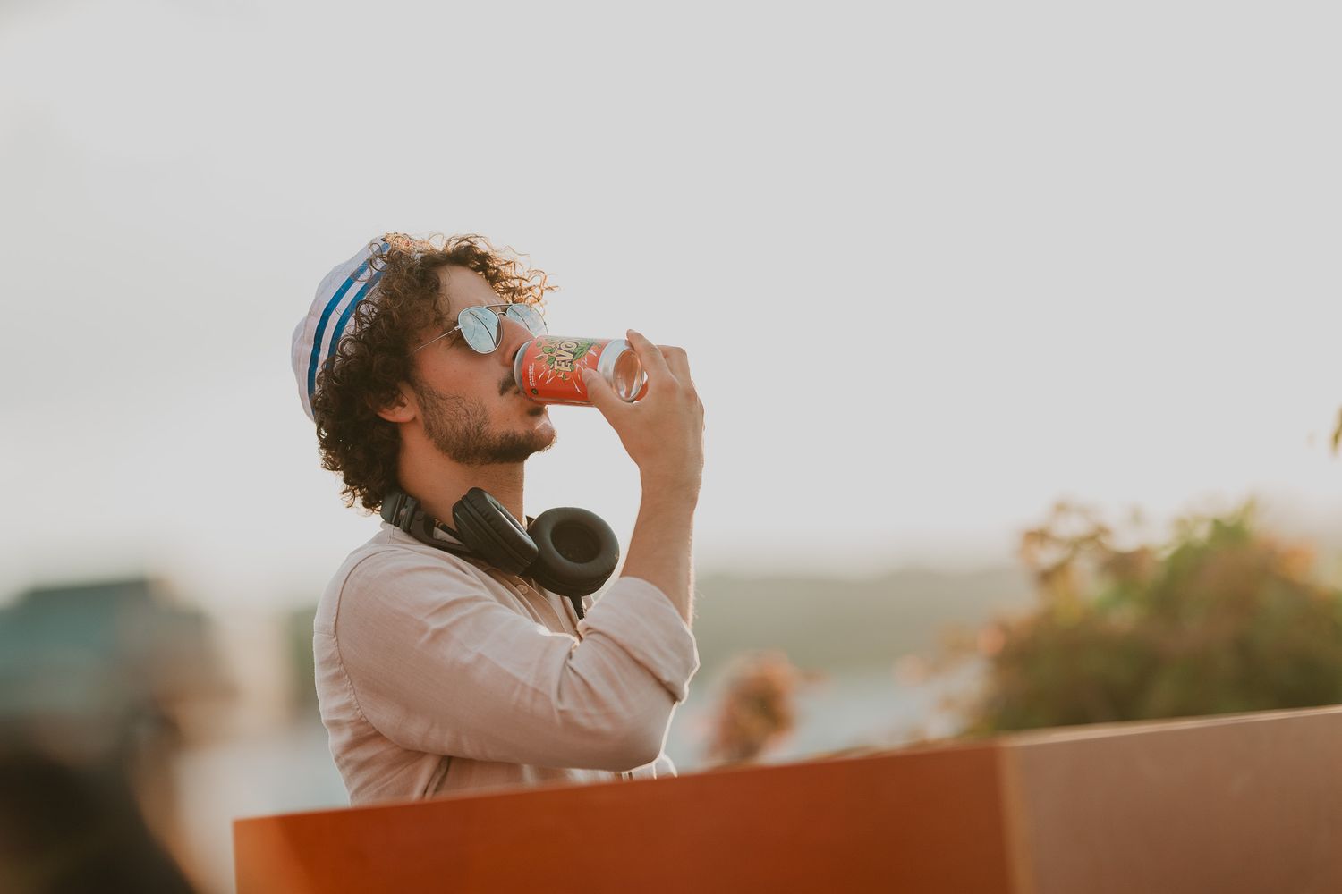 Young man with curly hair drinking from a can while wearing sunglasses outdoors.
