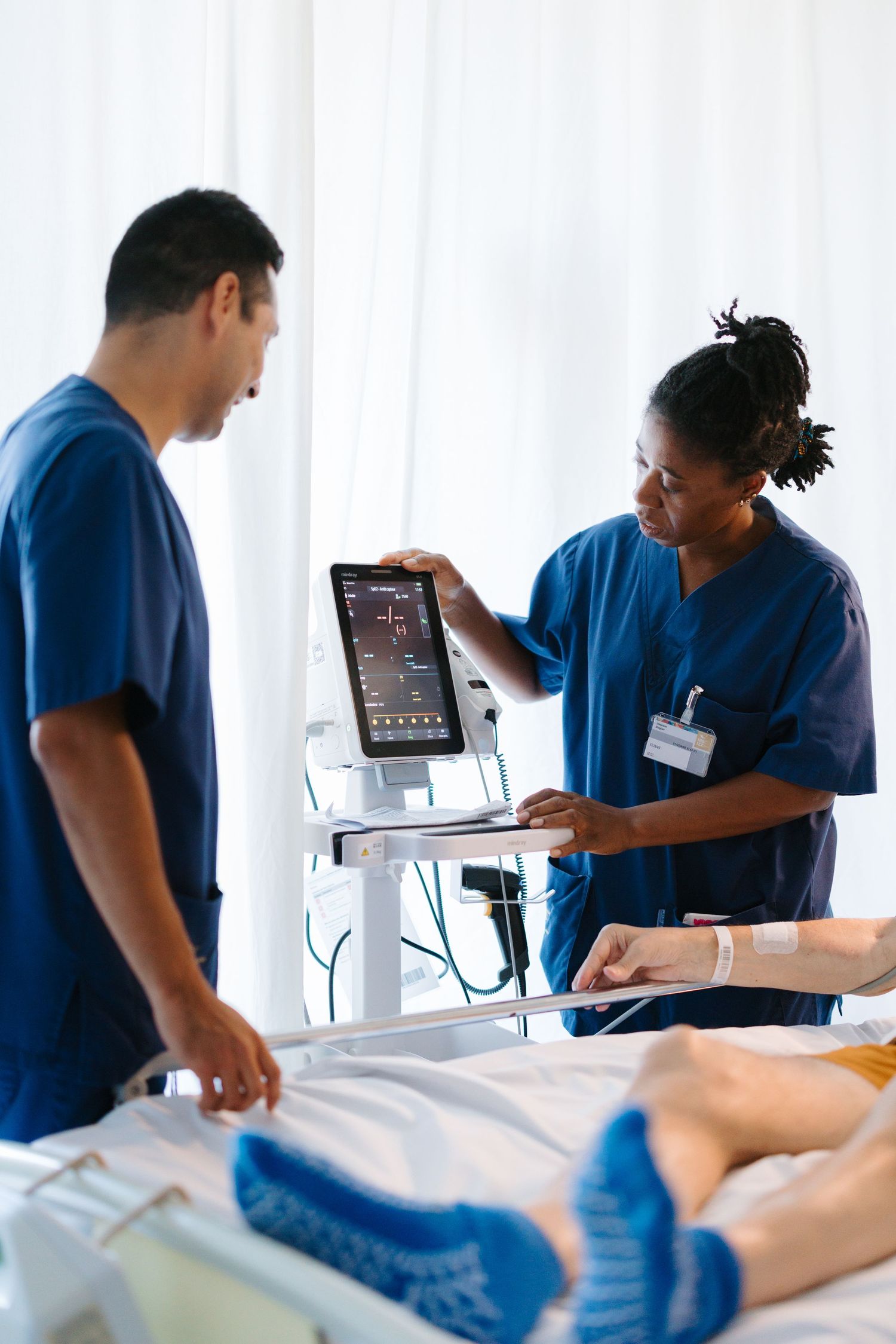 Two nurses in blue scrubs review patient monitor data at a hospital bedside during a medical consultation.