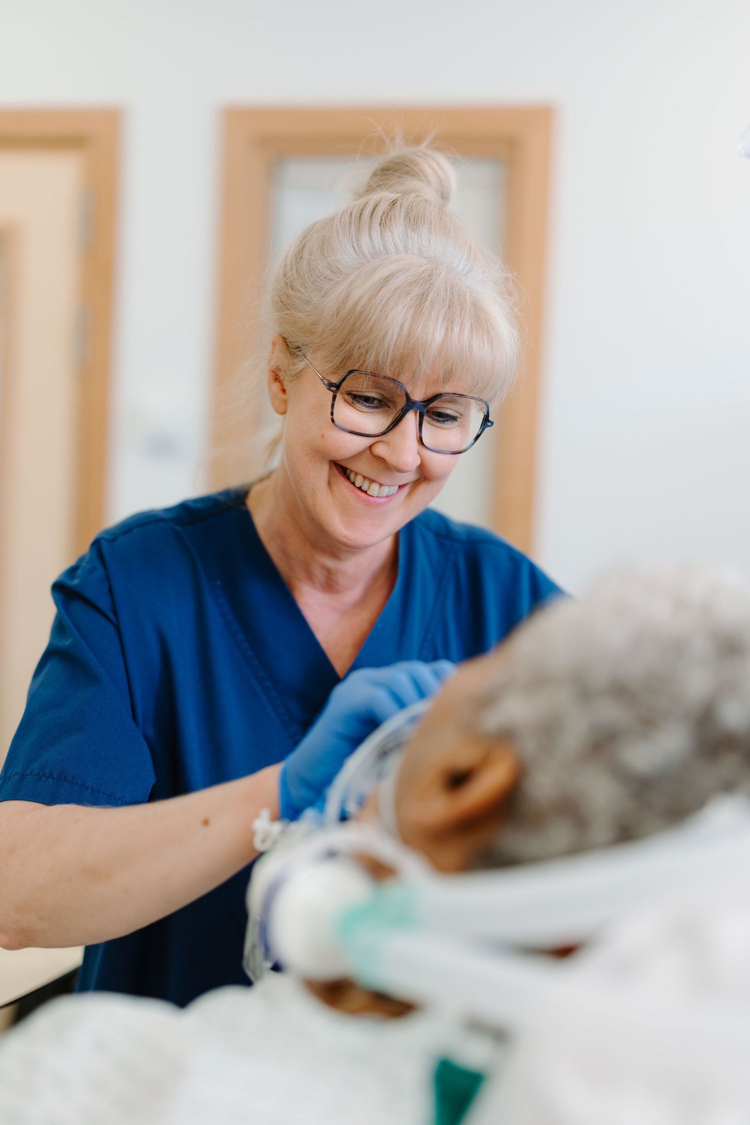 Smiling female nurse in blue scrubs and glasses providing care to a patient in a medical facility.