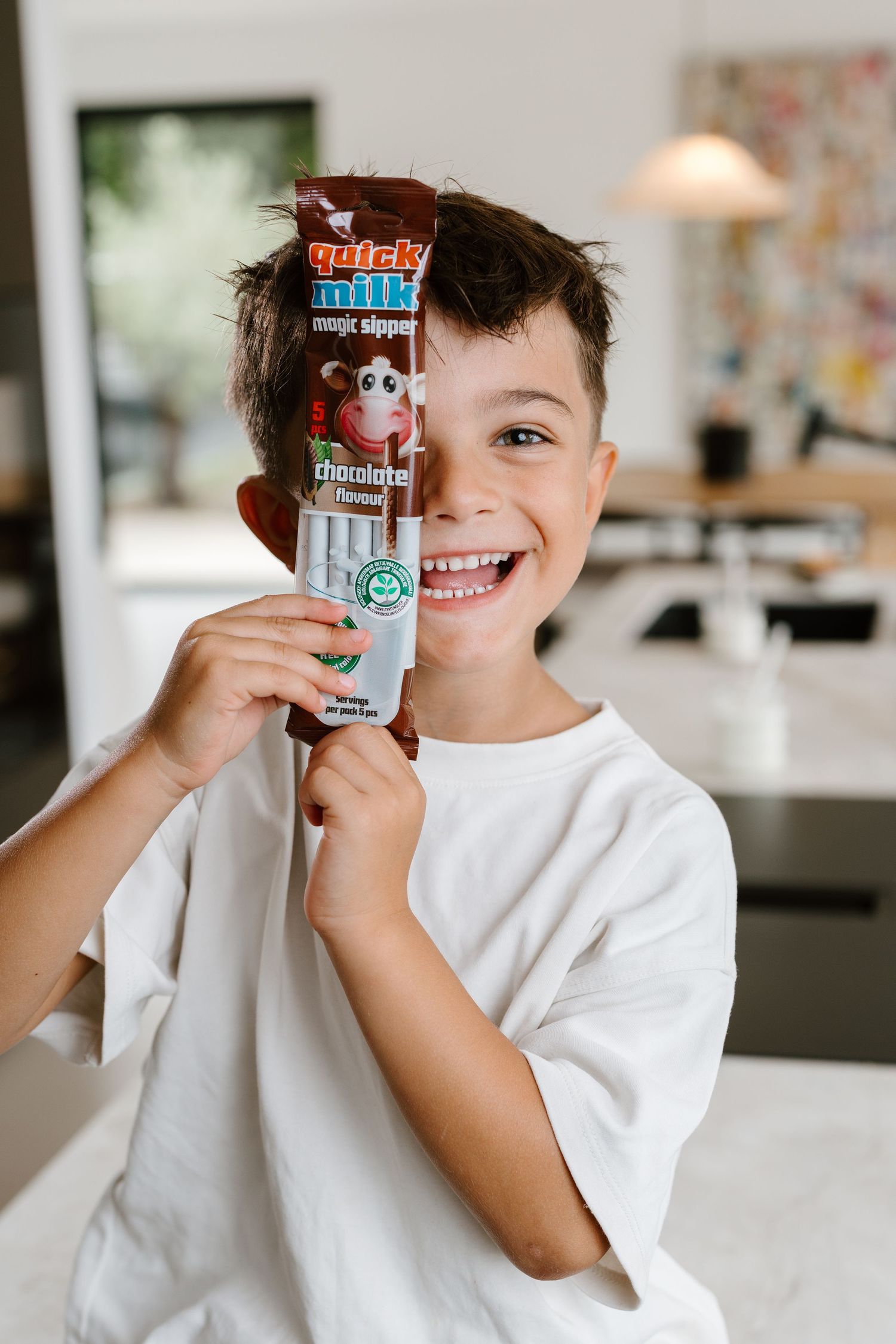 Smiling boy holding Quick Milk magic sipper chocolate flavored drink packet in a bright kitchen.