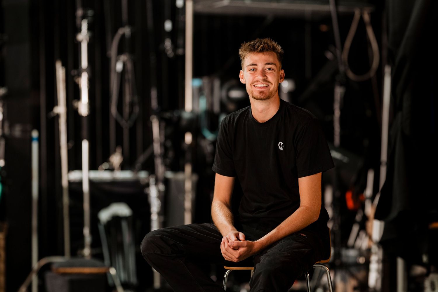 Young smiling man in black t-shirt sitting on chair in dark backstage theater environment.
