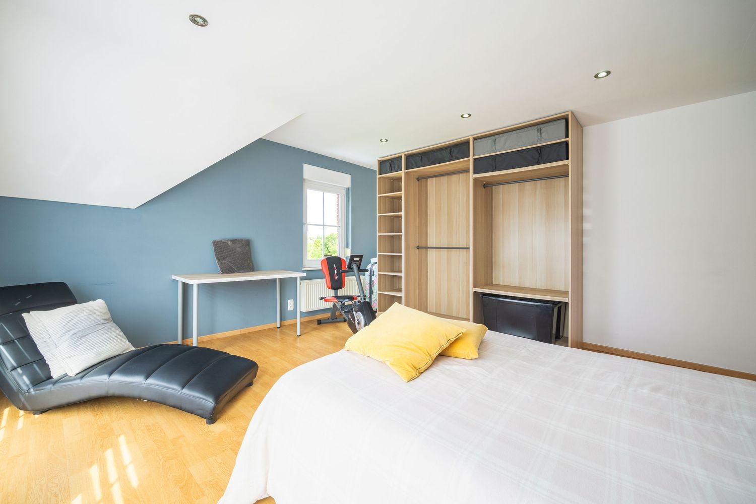 Bright attic bedroom with blue accent wall, built-in wooden wardrobe, white bed with yellow pillows, and a black lounge chair.