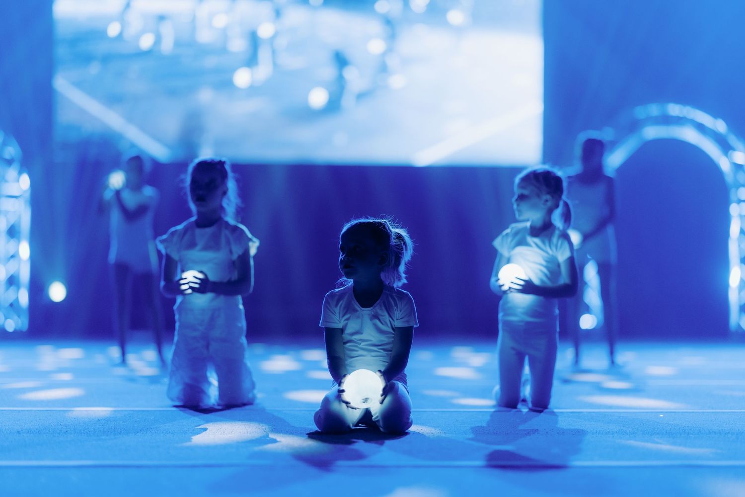 Children kneel on stage holding glowing orbs in a blue-lit theatrical performance.