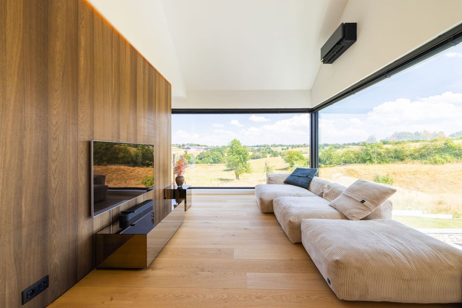 Modern minimalist living room with wood-paneled walls, white sofa, wall-mounted TV, and large panoramic windows overlooking green landscape.