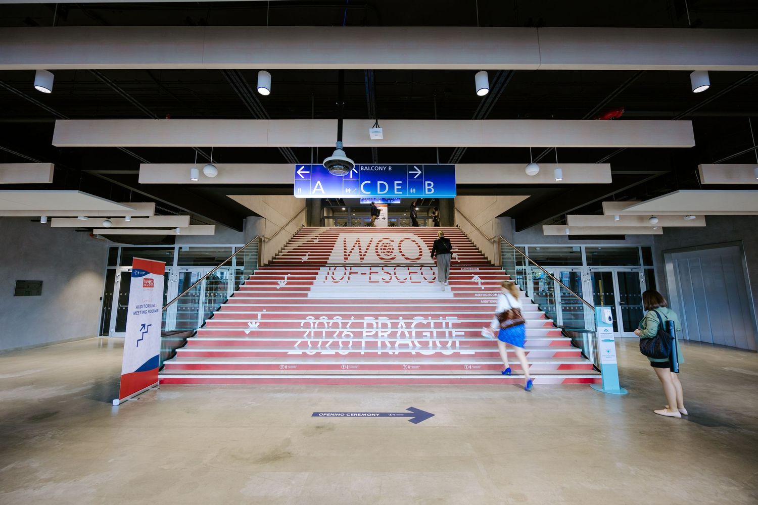 Grand stadium entrance with WCO branded stairs, digital scoreboard, and visitors walking toward glass doors.