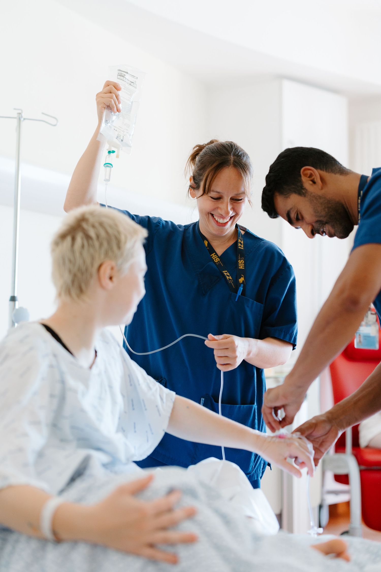 Two nurses in blue scrubs administering an IV drip to a young patient lying in a hospital bed.