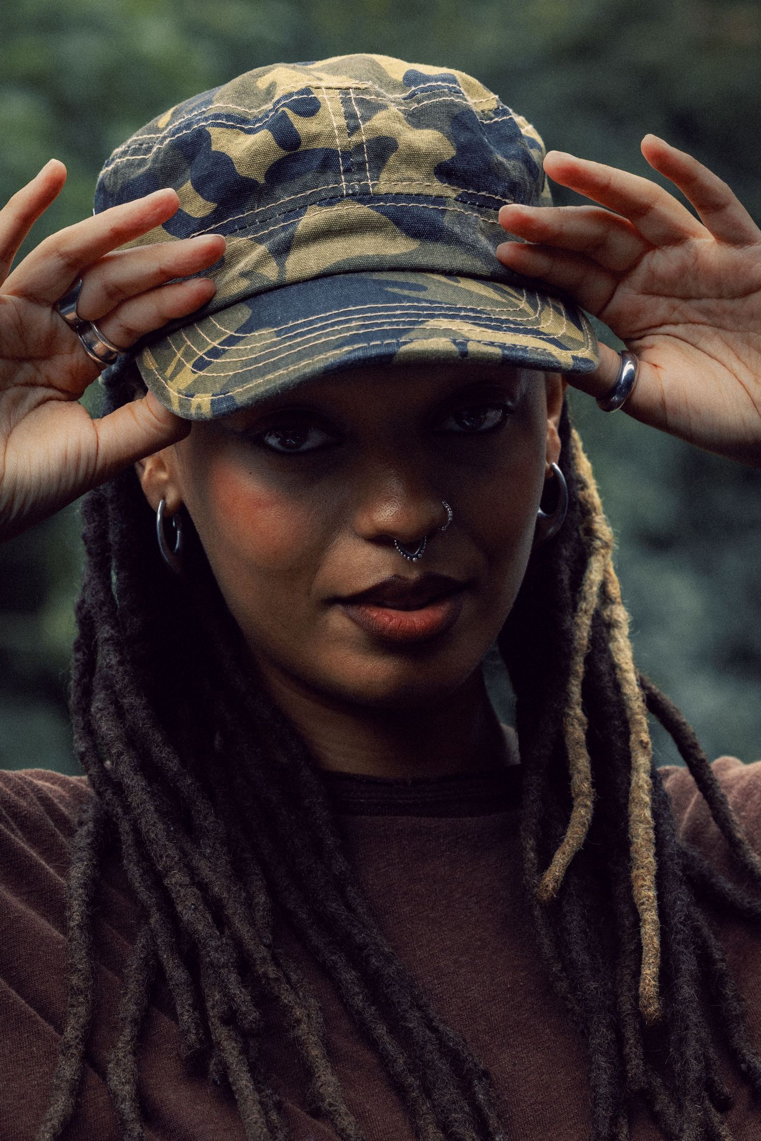 Young Black woman with dreadlocks wearing a camouflage cap and nose ring, posing outdoors.