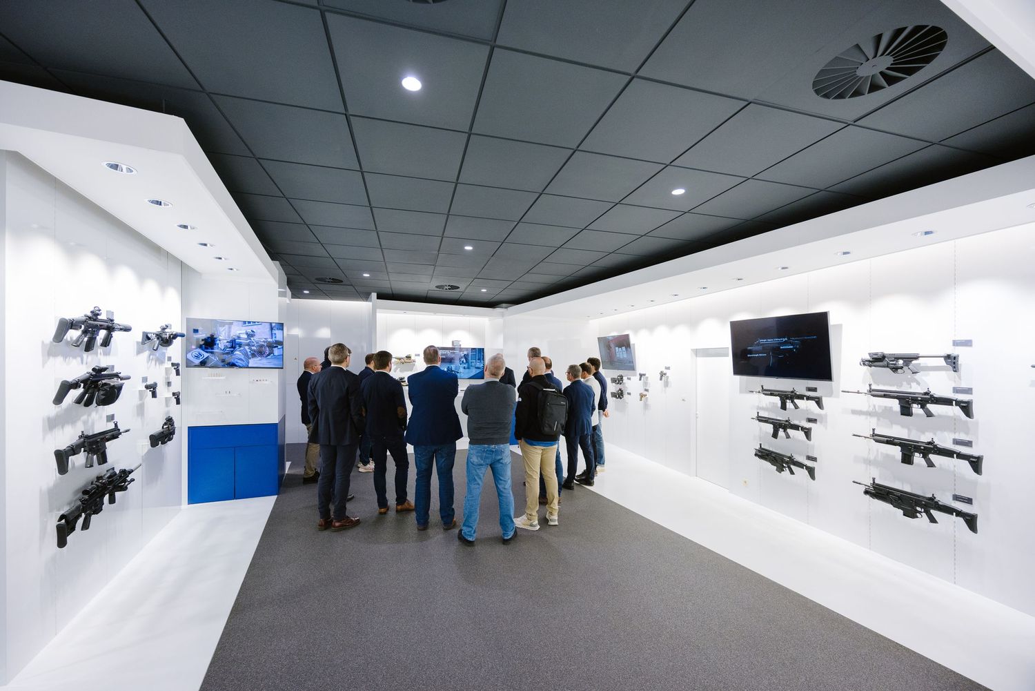 Group of visitors viewing firearms display at modern blue and white exhibition showroom.