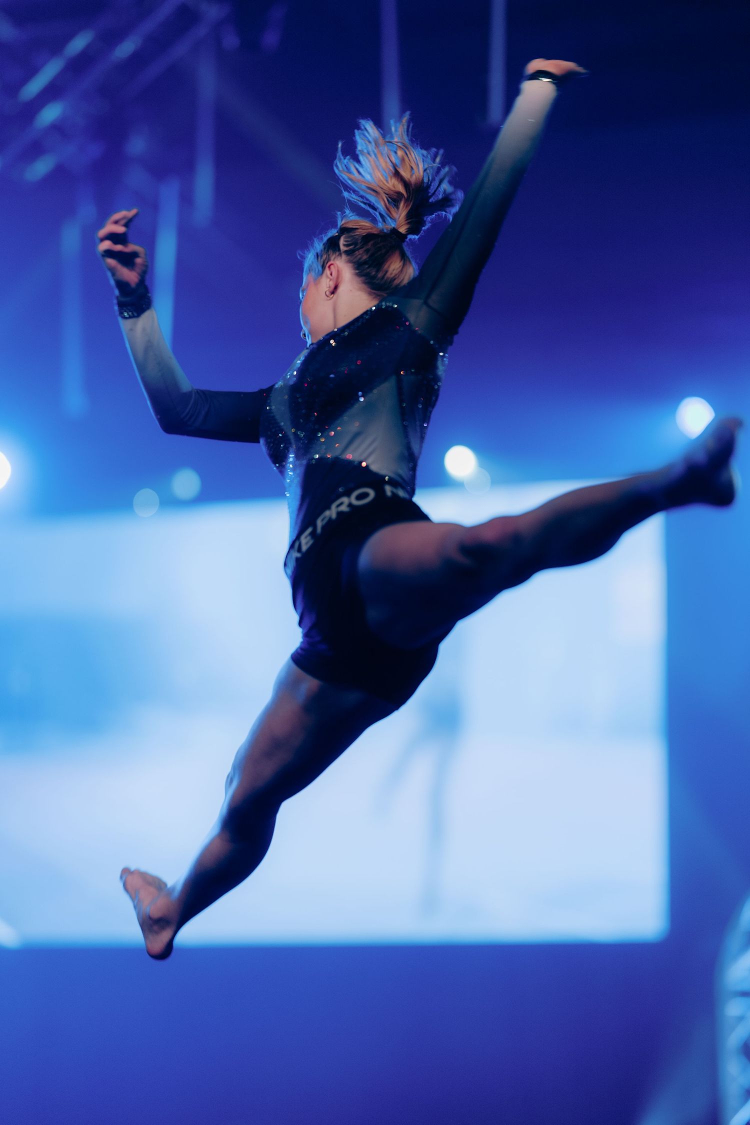 Gymnast leaps mid-air in a sparkling black leotard under blue stage lighting.