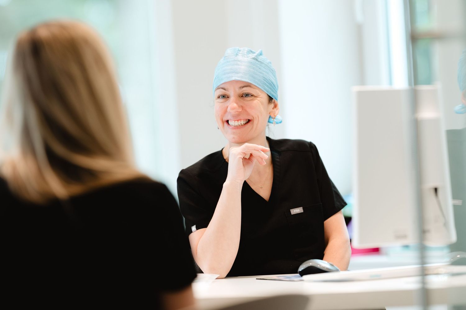 Smiling female surgeon in blue scrub cap and black scrubs consulting with a patient at a medical reception desk.