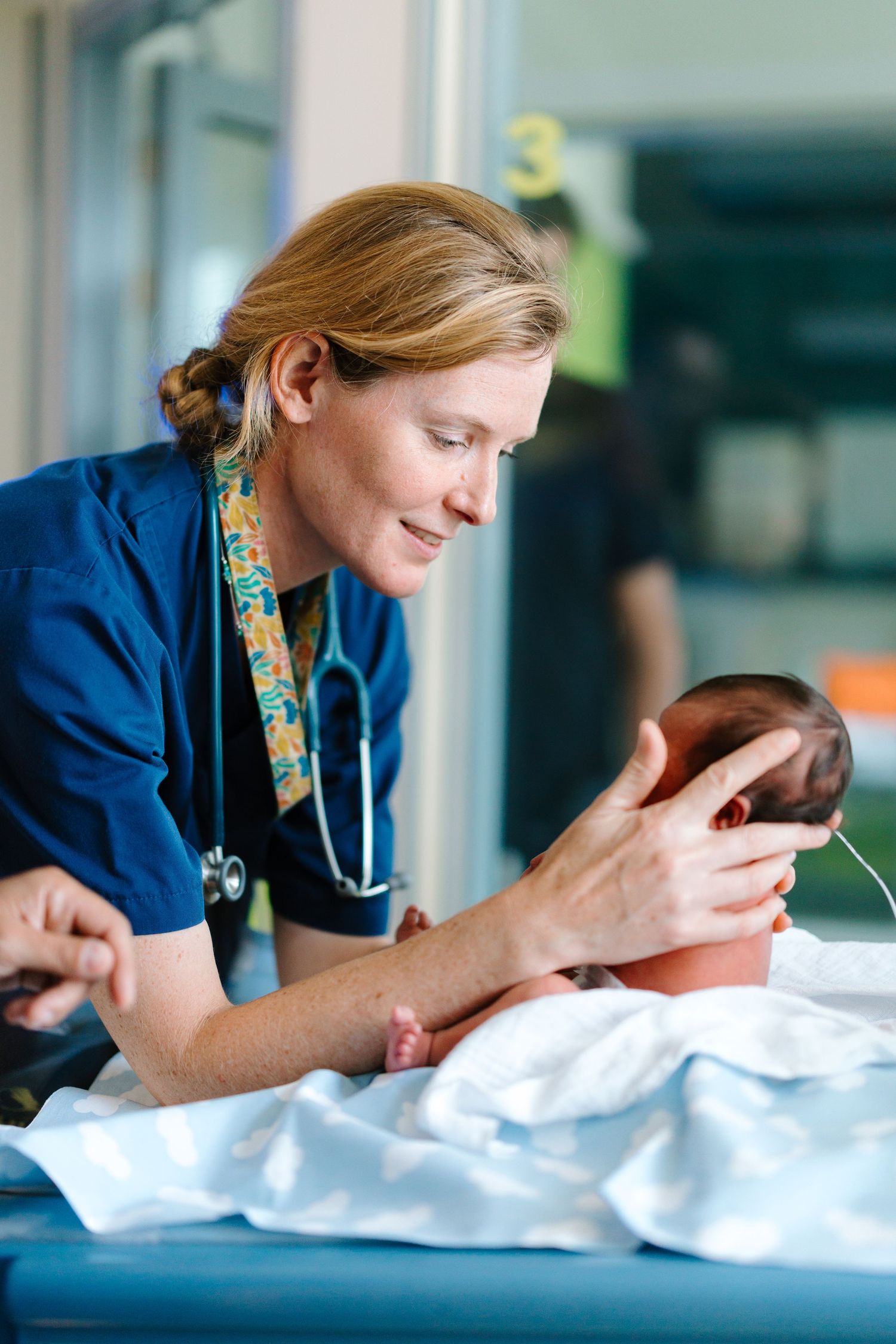 A smiling nurse in blue scrubs gently holds a newborn baby in a hospital setting.