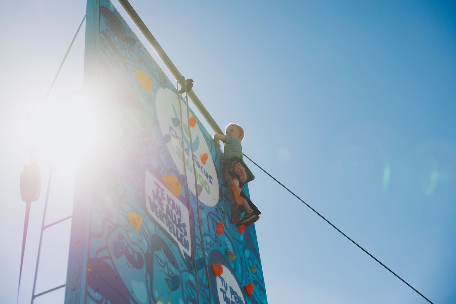 Child climbing colorful outdoor rock wall against bright blue sunny sky.