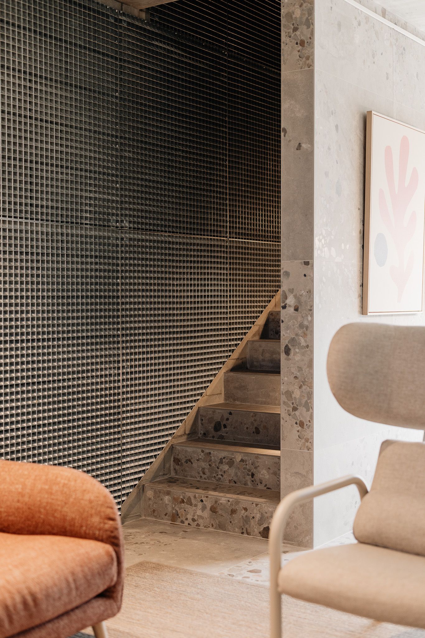 Modern interior staircase with dark grid mesh wall, terrazzo steps, and beige lounge chairs beside abstract wall art.