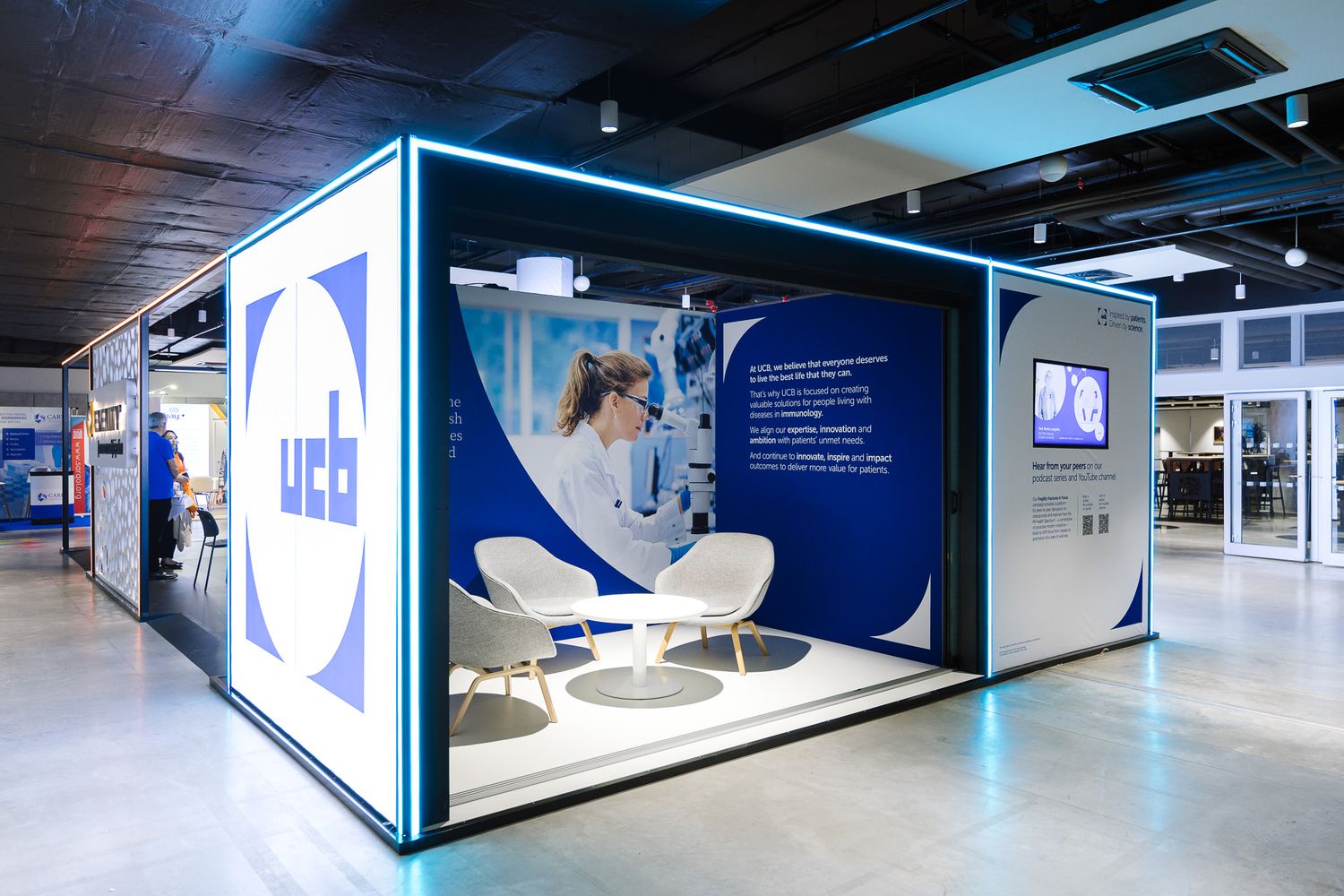 Modern UCB branded exhibition booth with white chairs and blue neon lighting at a trade show.