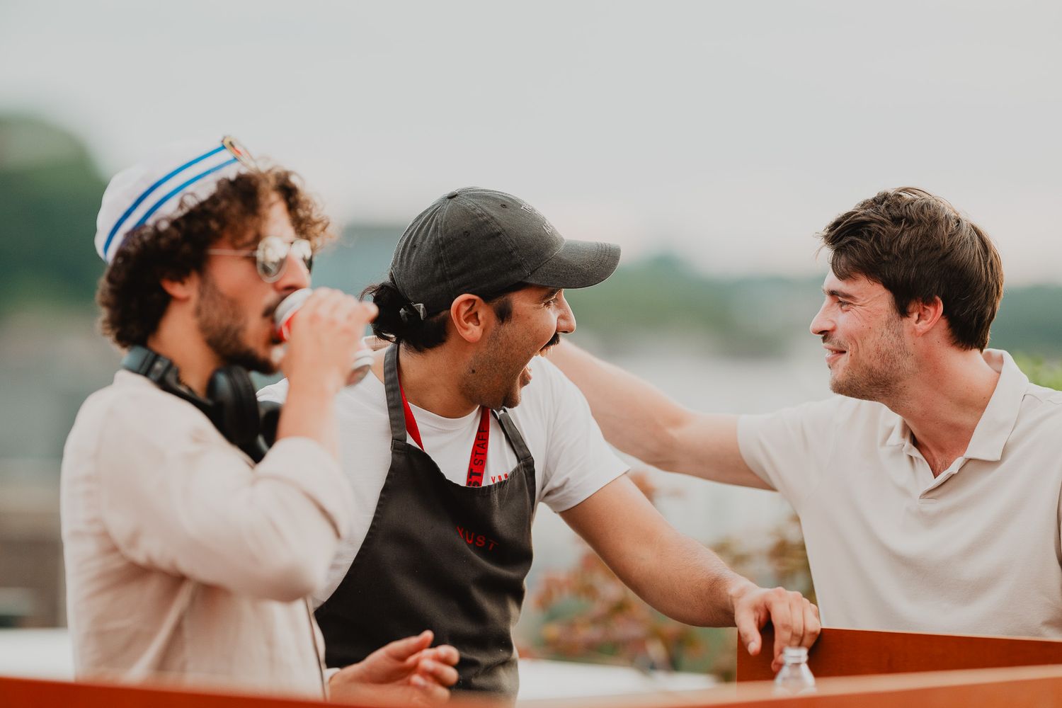 Three young men laughing and enjoying drinks together outdoors at a rooftop social gathering.