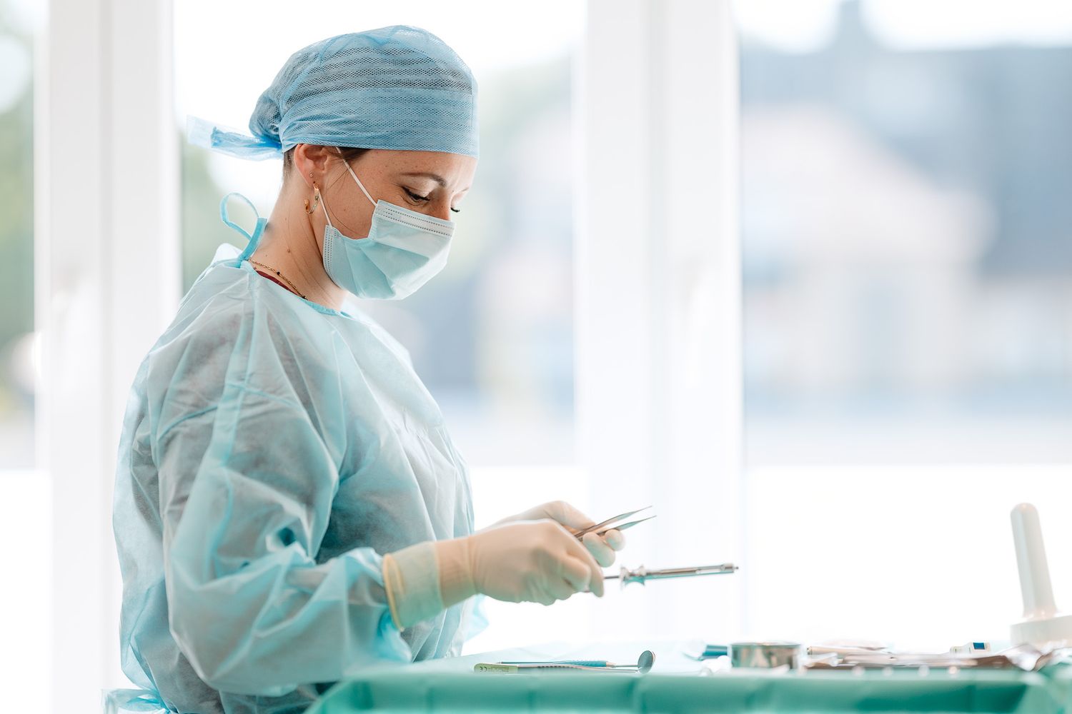 Female surgeon in blue scrubs and mask preparing medical instruments in bright operating room.
