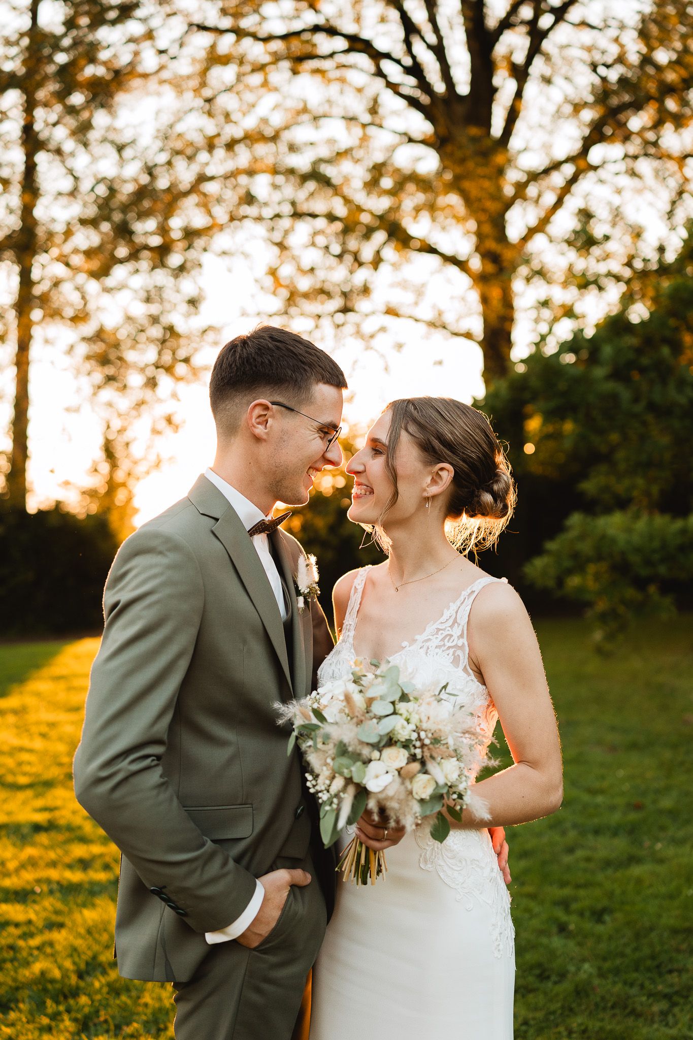 Smiling bride holding white bouquet with groom in green suit embracing at golden hour outdoors.