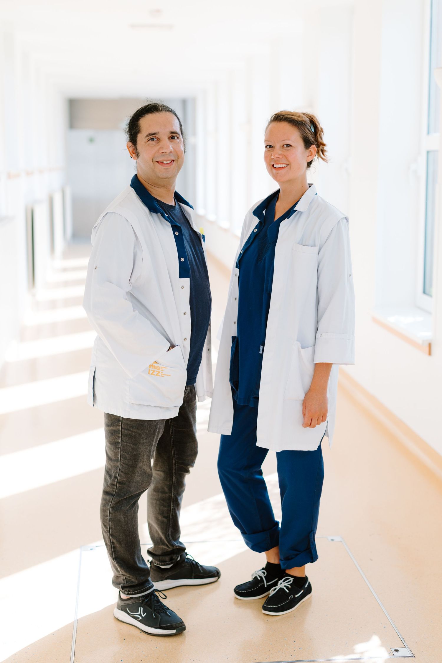 Two medical professionals in white lab coats and navy scrubs smiling in a bright hospital corridor.